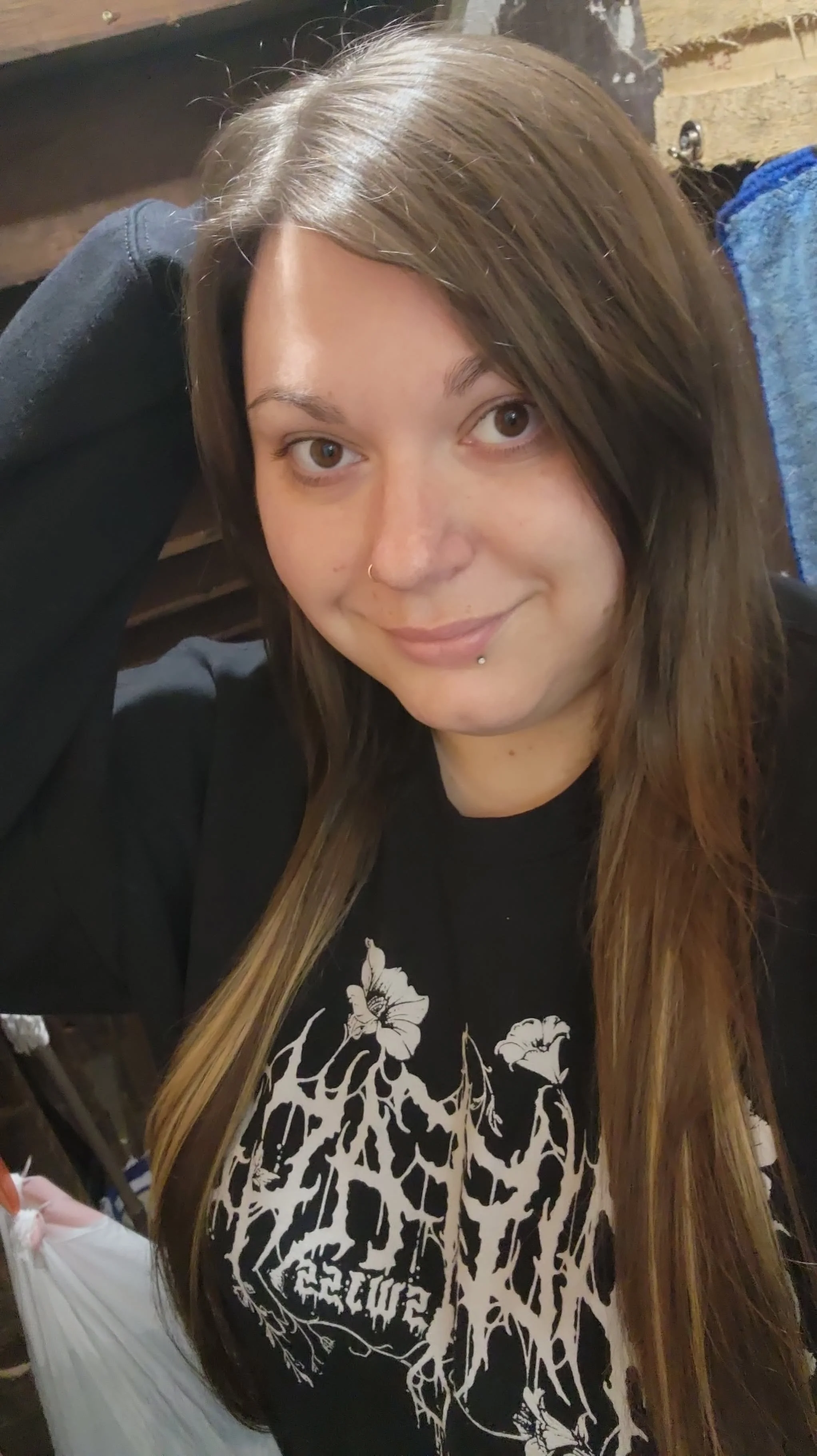 A young woman with long brown hair, a nose piercing, and a labret piercing, wearing a black graphic T-shirt with white floral and text design, smiling at the camera indoors.