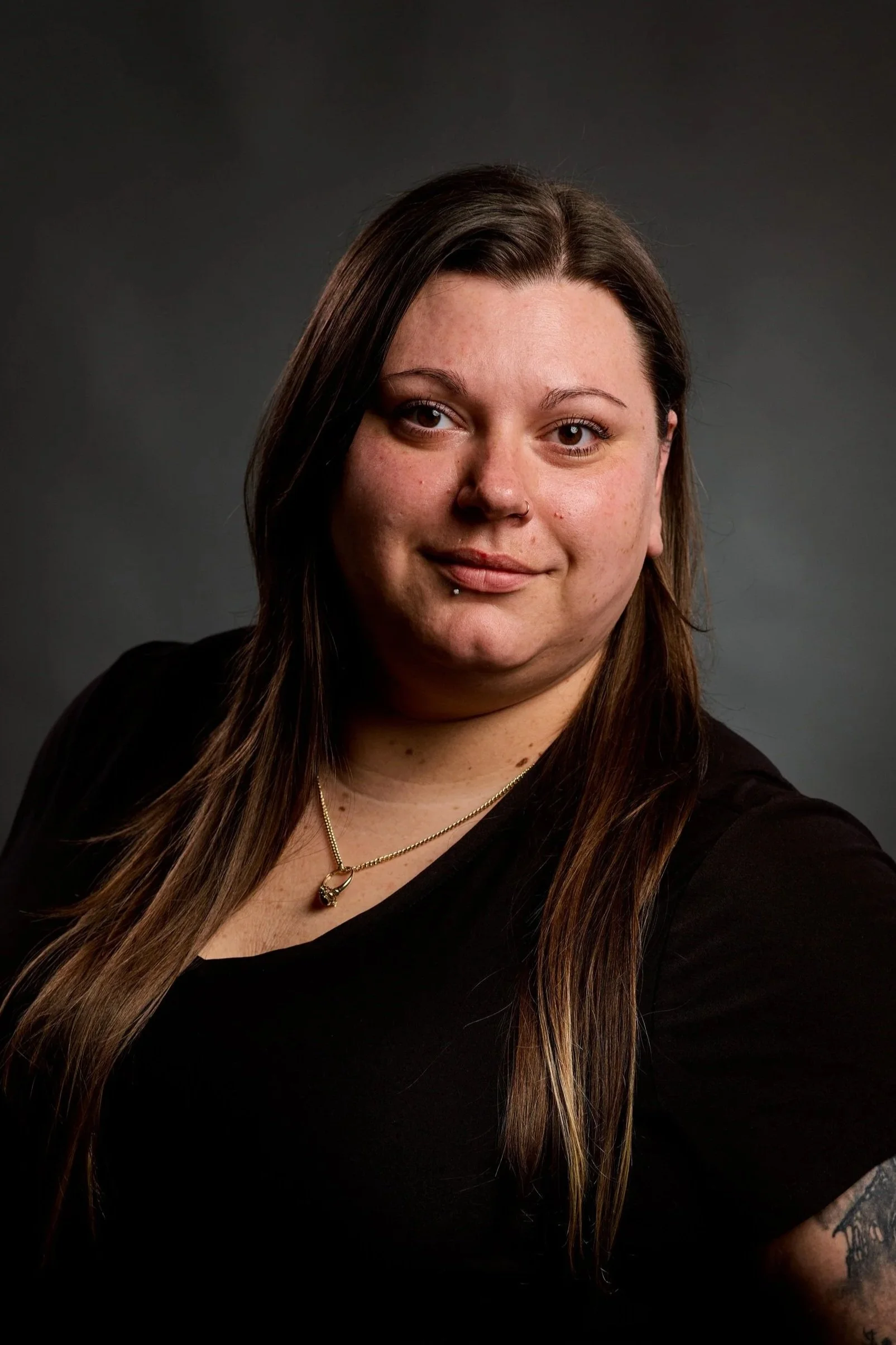 Portrait of a woman with long brown hair and a nose piercing, wearing a black top and gold jewelry, against a dark background.