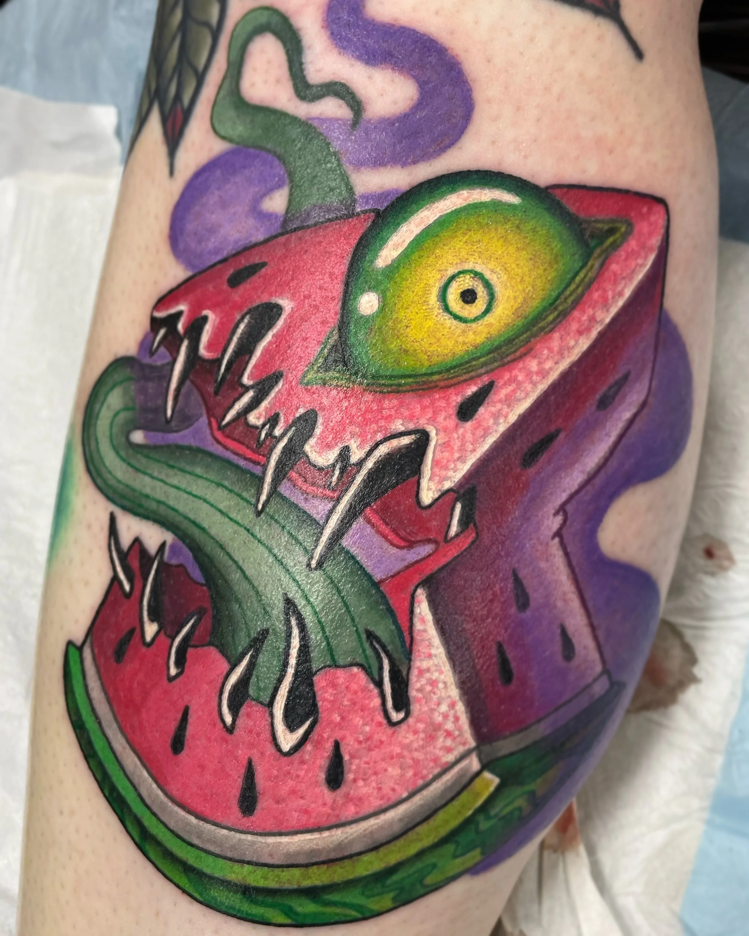 Colorful tattoo of a watermelon with a stylized eye inside it, on a person's upper arm.