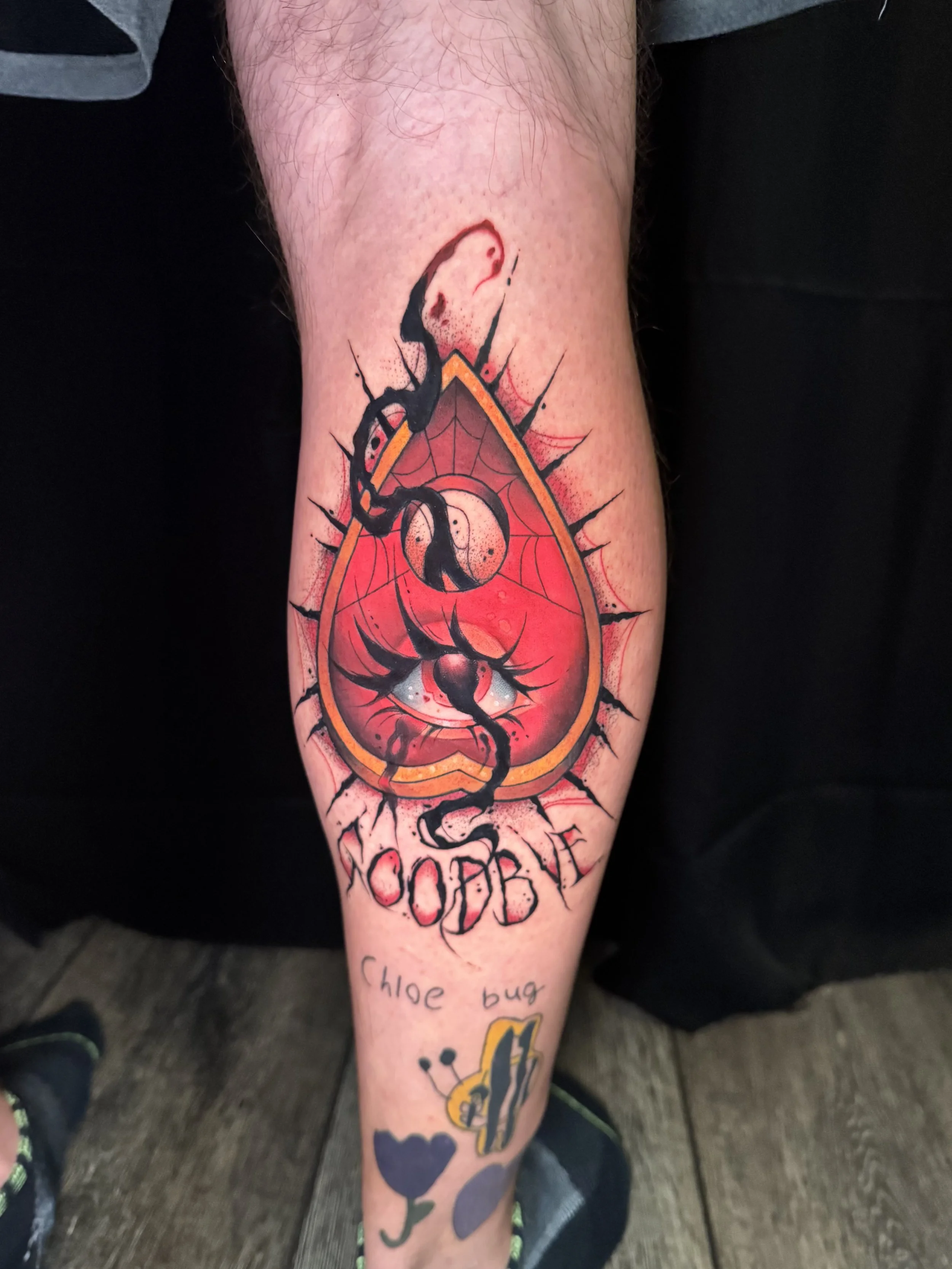 Tattoo on person's leg featuring a red and black design with a stylized insect, possibly a wasp, along with a blue heart and text that reads 'Chloe bug'.