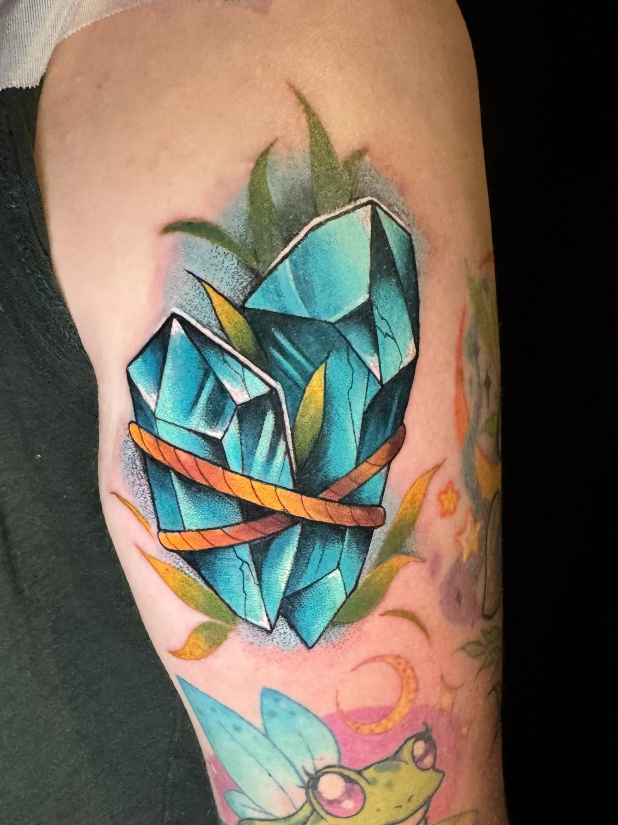 Colorful tattoo of a large blue diamond with green leaves and orange accents on a person's arm.