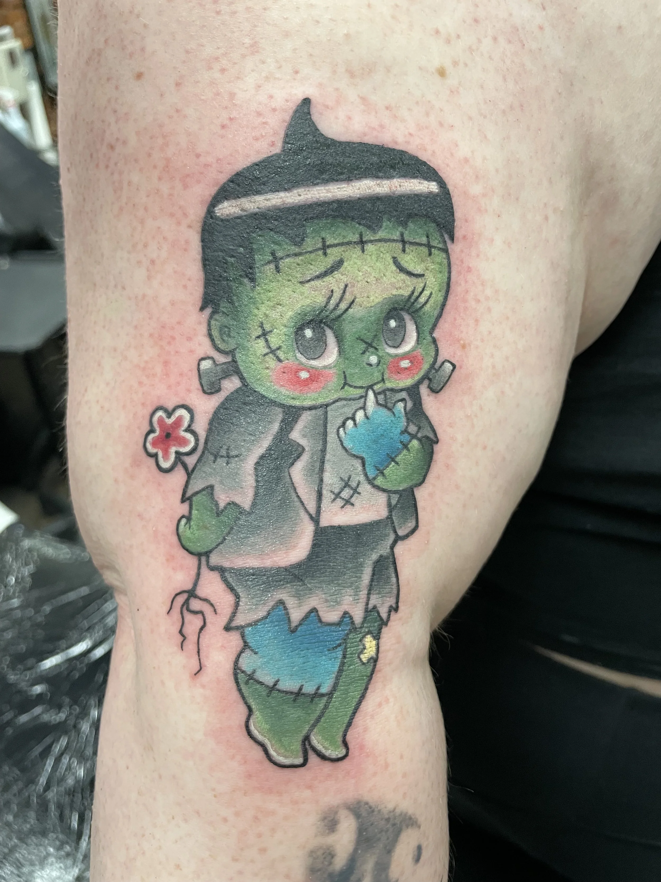 A colorful tattoo of a cartoon Frankenstein's monster with large eyes, green skin, and stitches, holding a blue heart, on a person's upper arm.