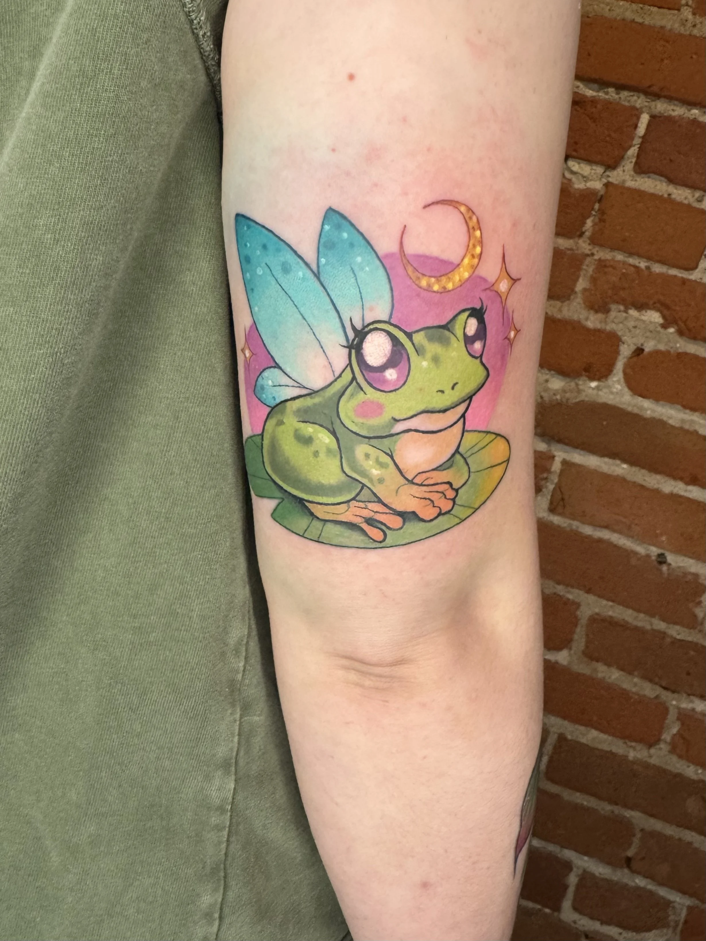 Colorful tattoo of a cute frog with fairy wings, a crescent moon, and sparkling stars on a person's upper arm.