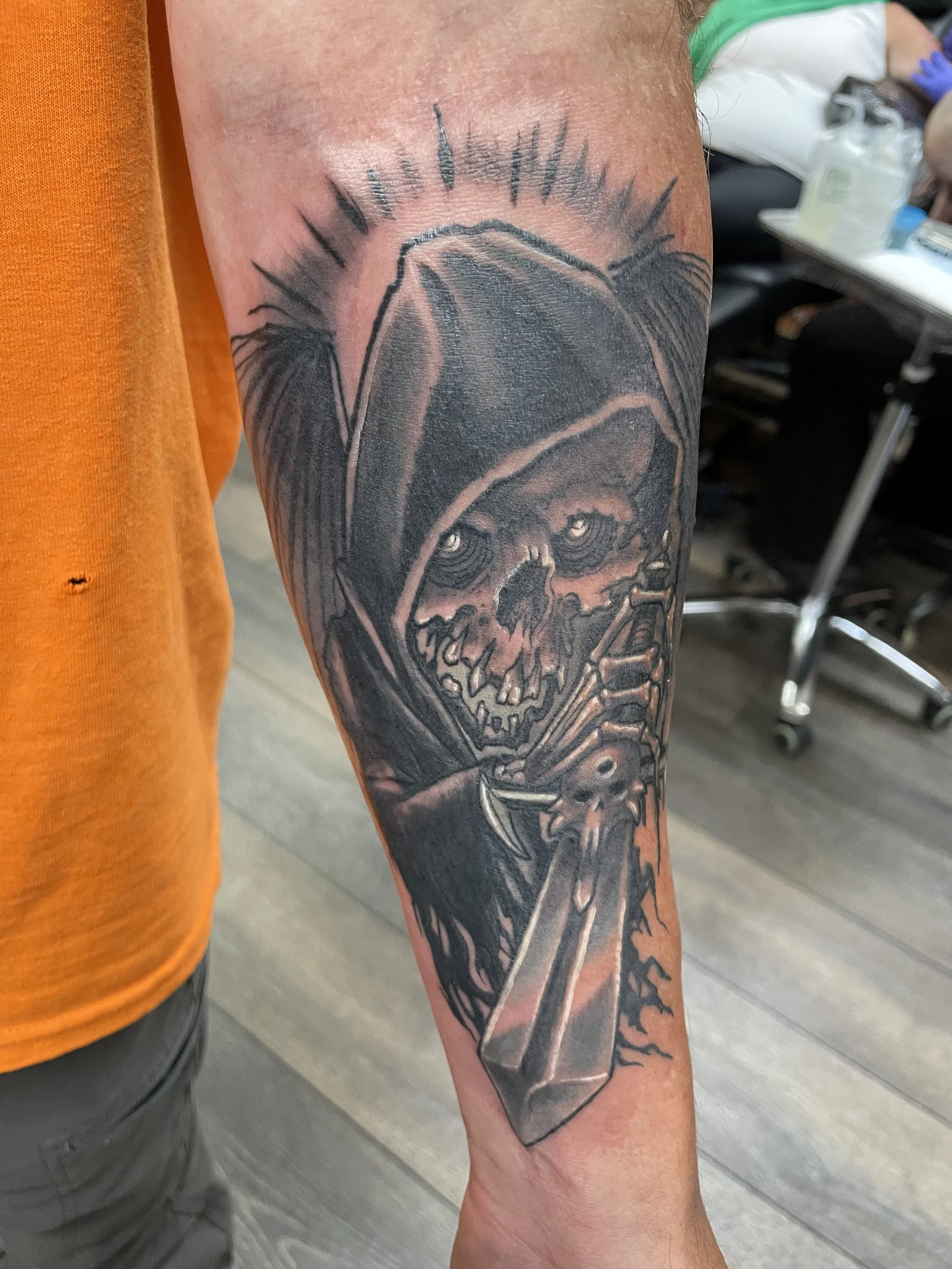 A tattoo of a human skull with sharp teeth on a person's forearm, featuring dark shading and outlined in black ink, with a stylized tree branch or lightning bolt extending from the skull.