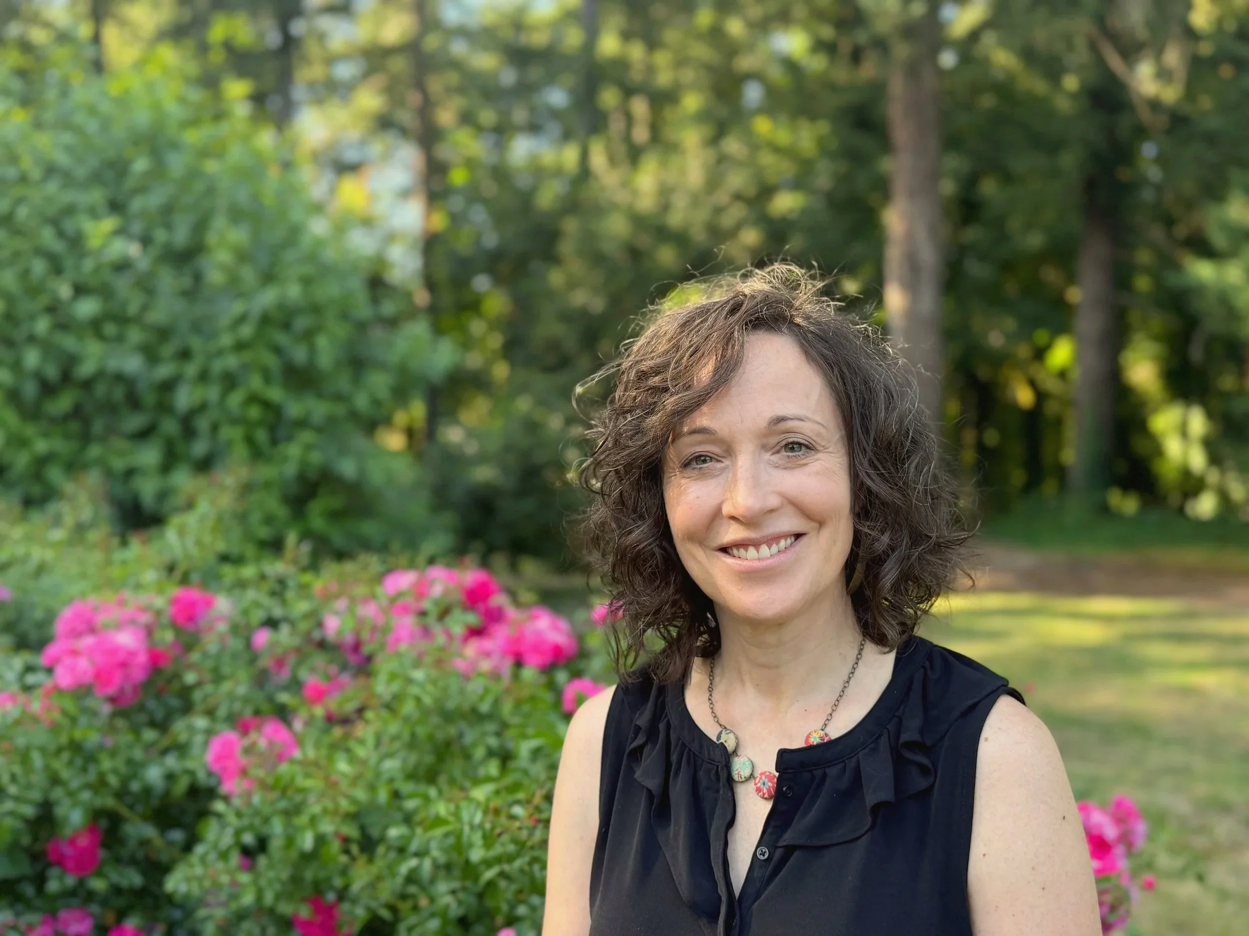 A smiling woman with curly brown hair and blue eyes, wearing a black sleeveless top and a colorful beaded necklace, standing outdoors in a park with green trees and pink flowers in the background.