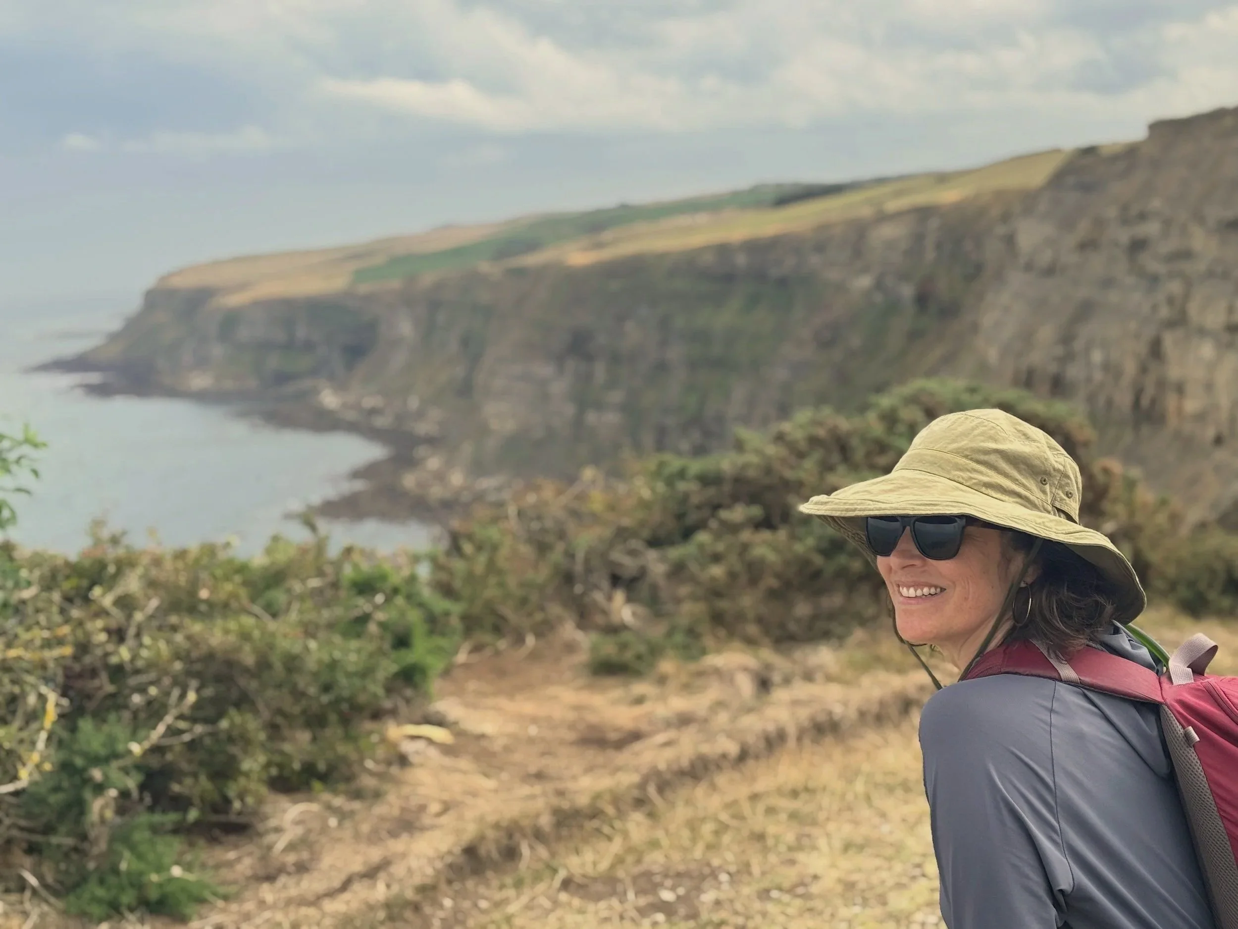 A woman wearing a wide-brimmed hat, sunglasses, and a backpack, smiling while walking outdoors near coastal cliffs.