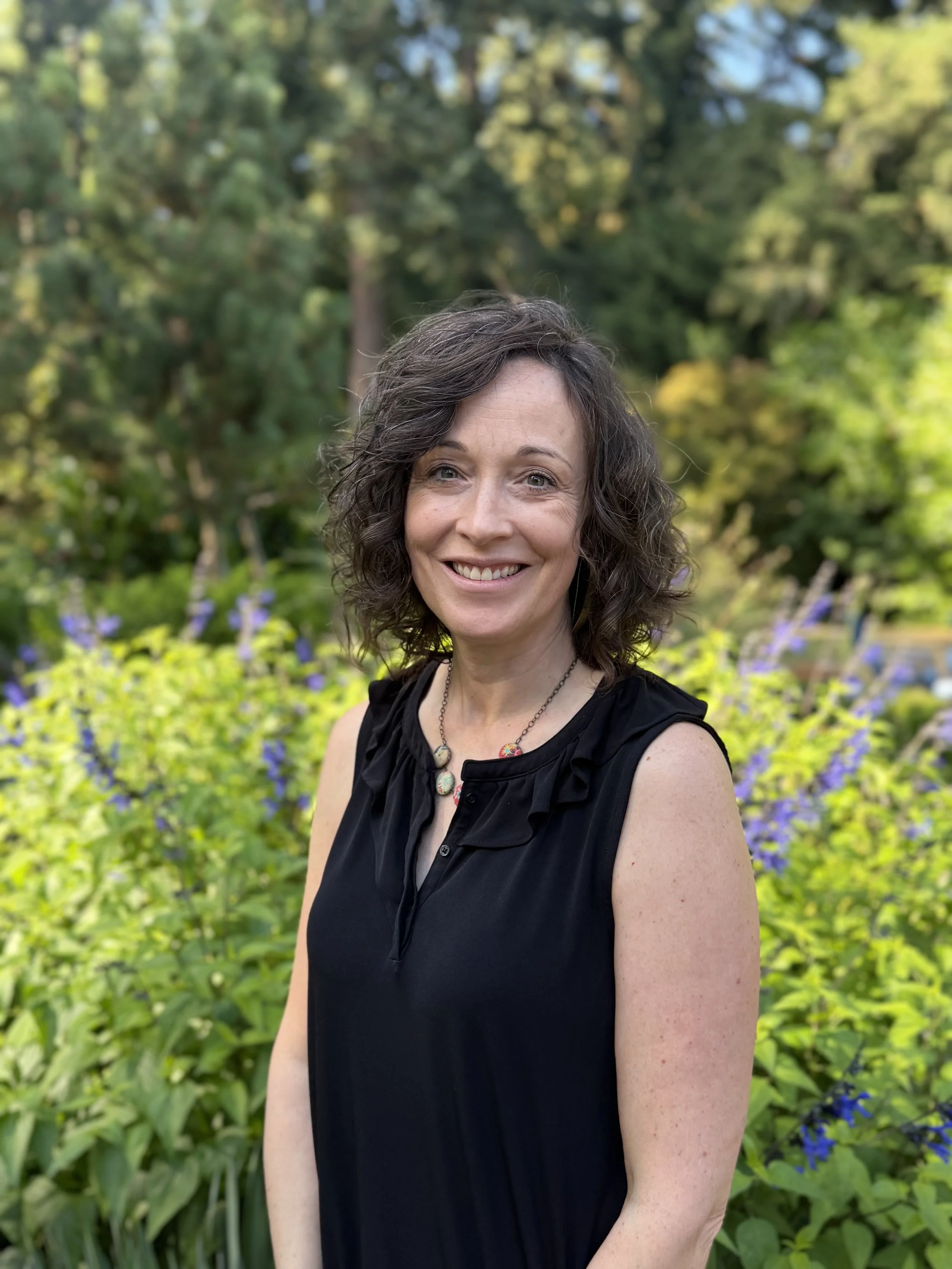A smiling woman with shoulder-length curly brown hair, wearing a black sleeveless top and a colorful beaded necklace, standing outdoors in a garden with green foliage and purple flowers in the background.