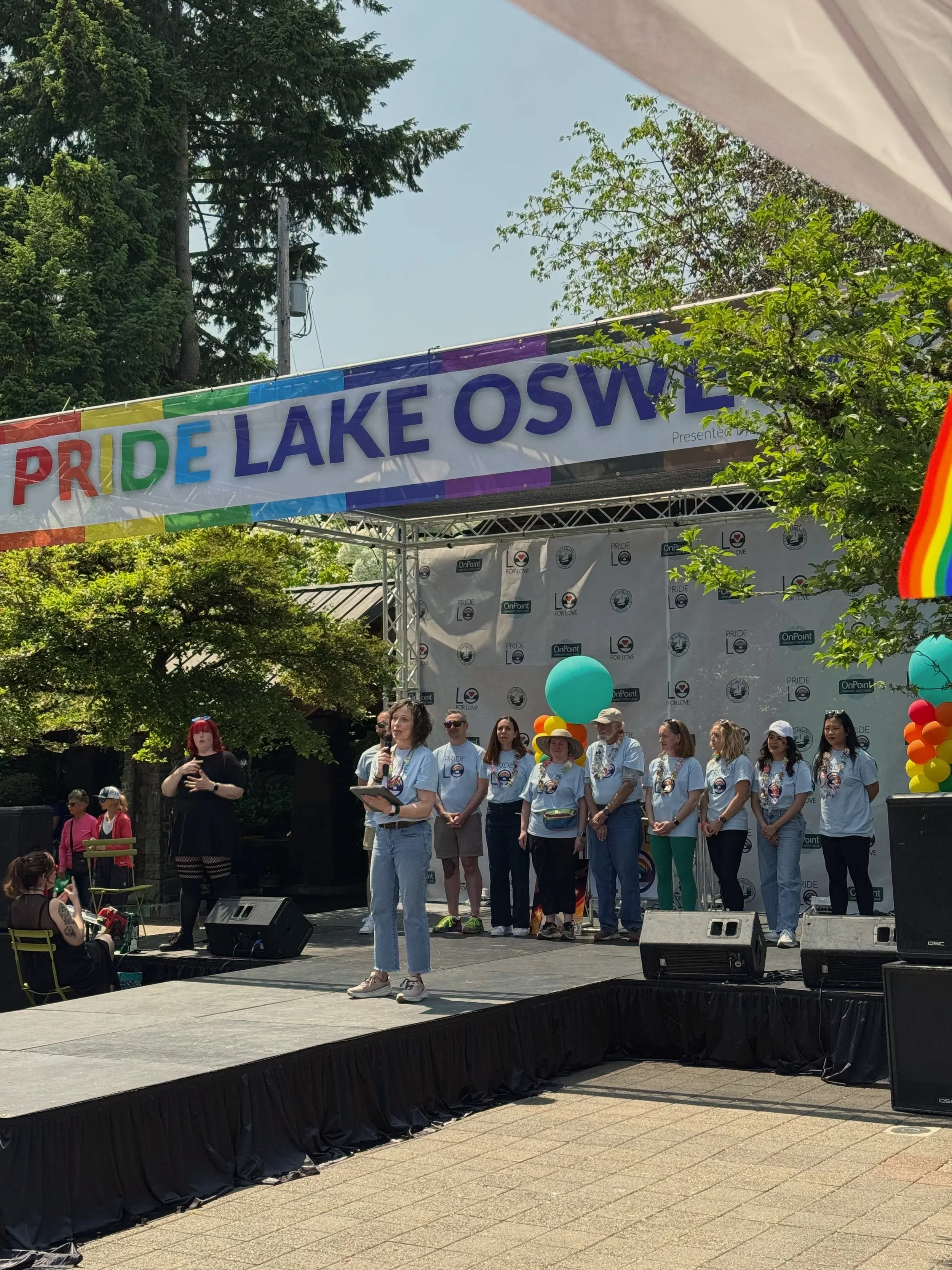 People gathered on an outdoor stage at a pride event, with a rainbow-themed banner reading 'PRIDE LAKE OSWEGO' overhead. Some individuals are wearing T-shirts with pride logos, and one woman is speaking into a microphone. There are colorful balloons and trees in the background, indicating a festive celebration.