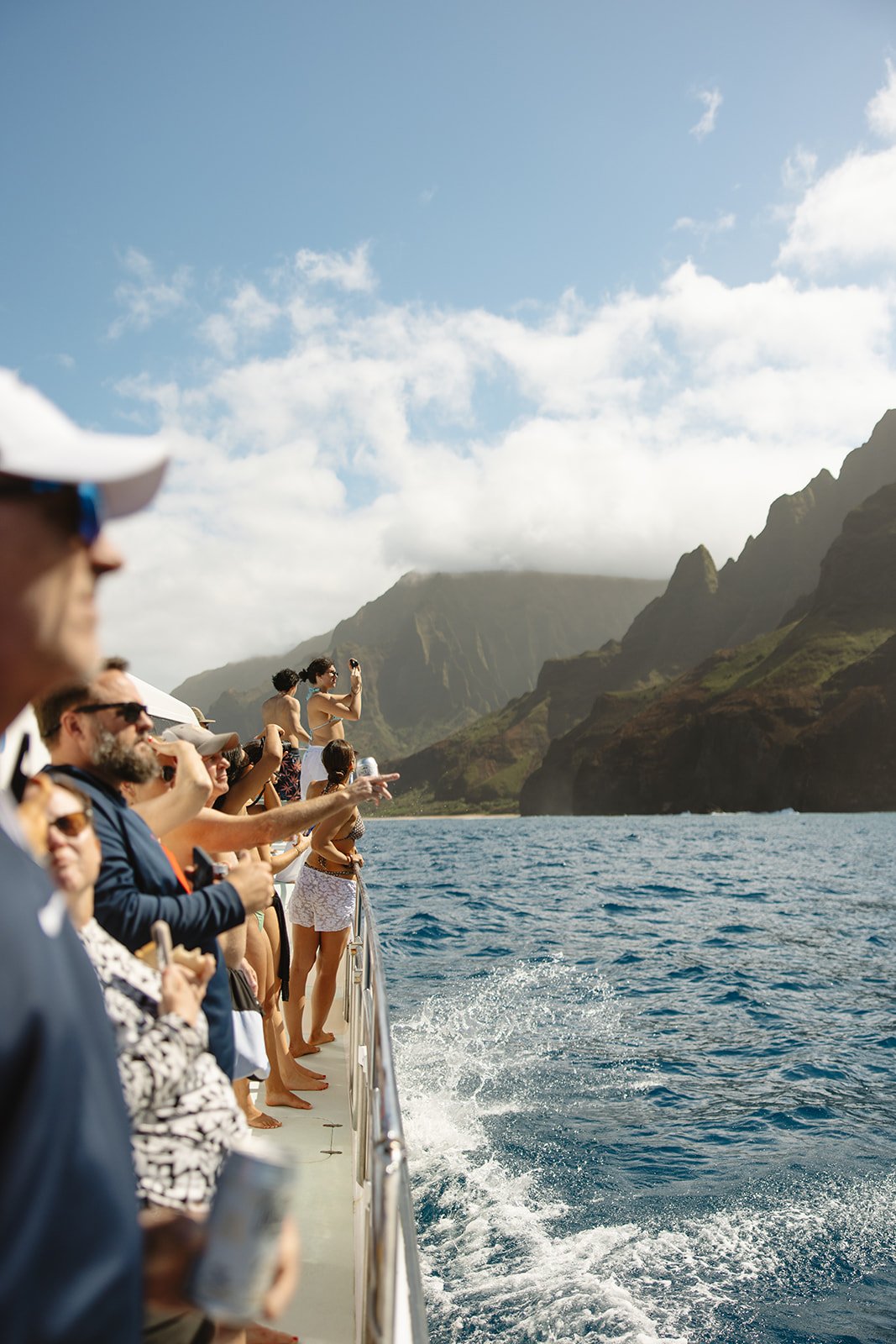 People on a boat cruise enjoying scenic mountain views and ocean, some taking photos, with a partly cloudy sky overhead. Brand and travel photography session for Holo Holo Charters in Kauai, Hawaii by Rhianon Brown Photography.