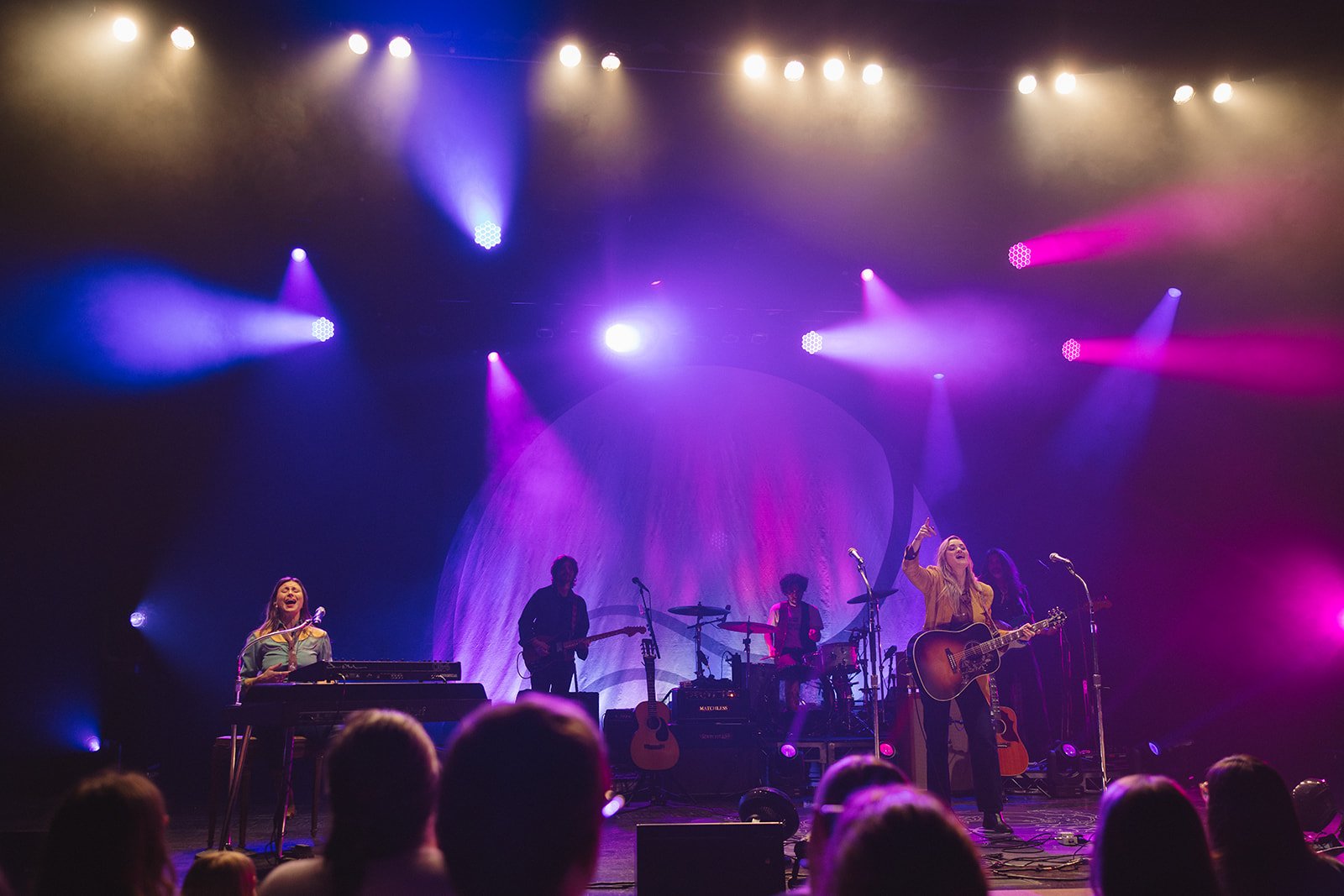A band performing on stage with colorful lights, including a woman playing guitar and singing, a woman playing keyboard, and three other band members with guitars, all under purple and pink stage lighting. Aly and Aj concert event photographed by Rhi