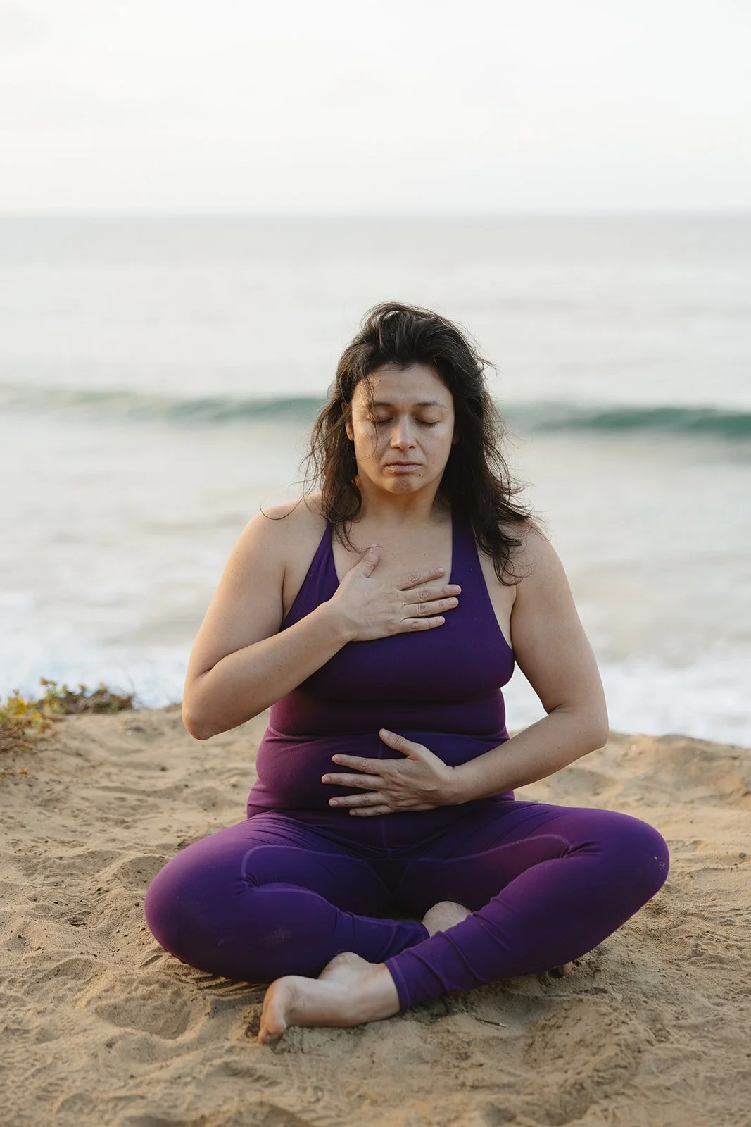 A woman sitting cross-legged on the sand at the beach, practicing meditation or yoga with her eyes closed. Brand and lifestyle photography for Connie Neyra and yoga practitioner by Rhianon Brown Photography.