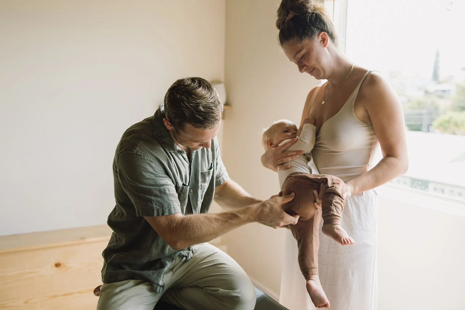 A woman holding a baby in one arm while breastfeeding, with a man adjusting the baby's diaper. Brand and lifestyle photography session for Abundant Life Chiropractics in Maui, Hawaii by Rhianon Brown Photography.