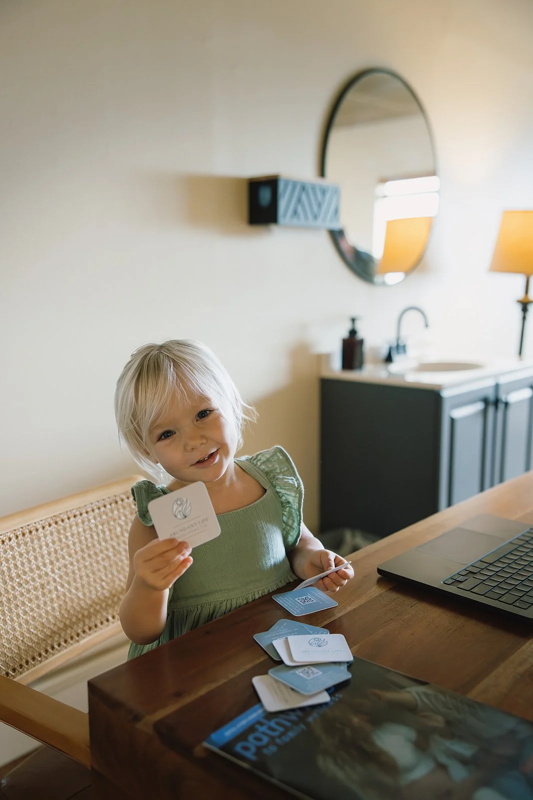A young girl with blonde hair sitting at a wooden table, holding a card, with a laptop and several other cards in front of her, smiling at the camera. Brand and lifestyle photography session for Abundant Life Chiropractics in Maui, Hawaii by Rhianon