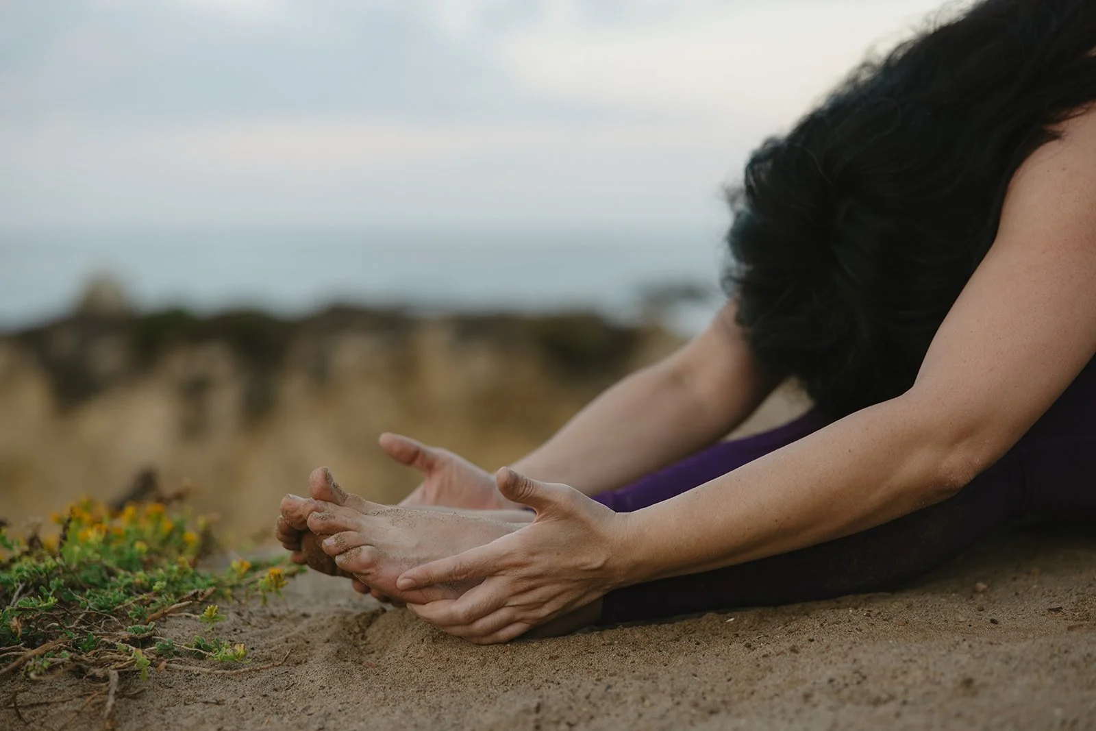 Person practicing yoga outdoors on sandy ground near grass and flowers, stretching forward with legs extended and hands reaching toward their feet. Brand and lifestyle photography for Connie Neyra and yoga practitioner by Rhianon Brown Photography.
