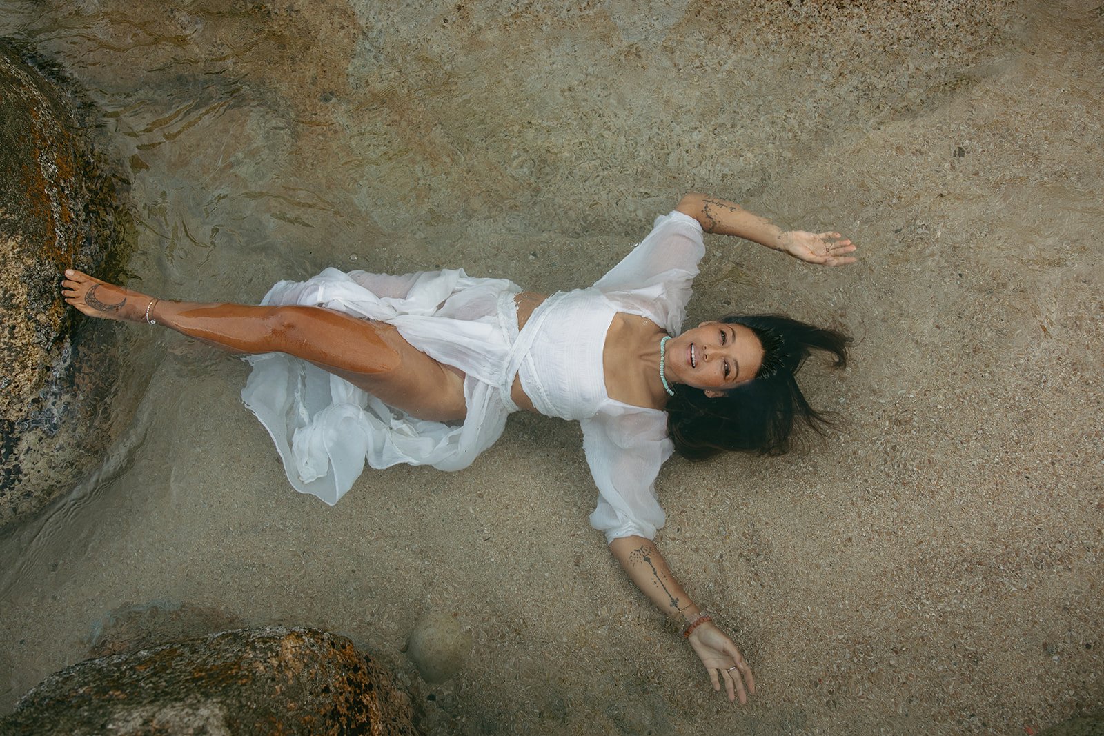 Woman lying on sandy beach with water and rocks, wearing white dress, smiling, with dark hair spread out, jewelry, and tattoos visible. Brand and retreat photography for Nikki Nie by Rhianon Brown Photography.