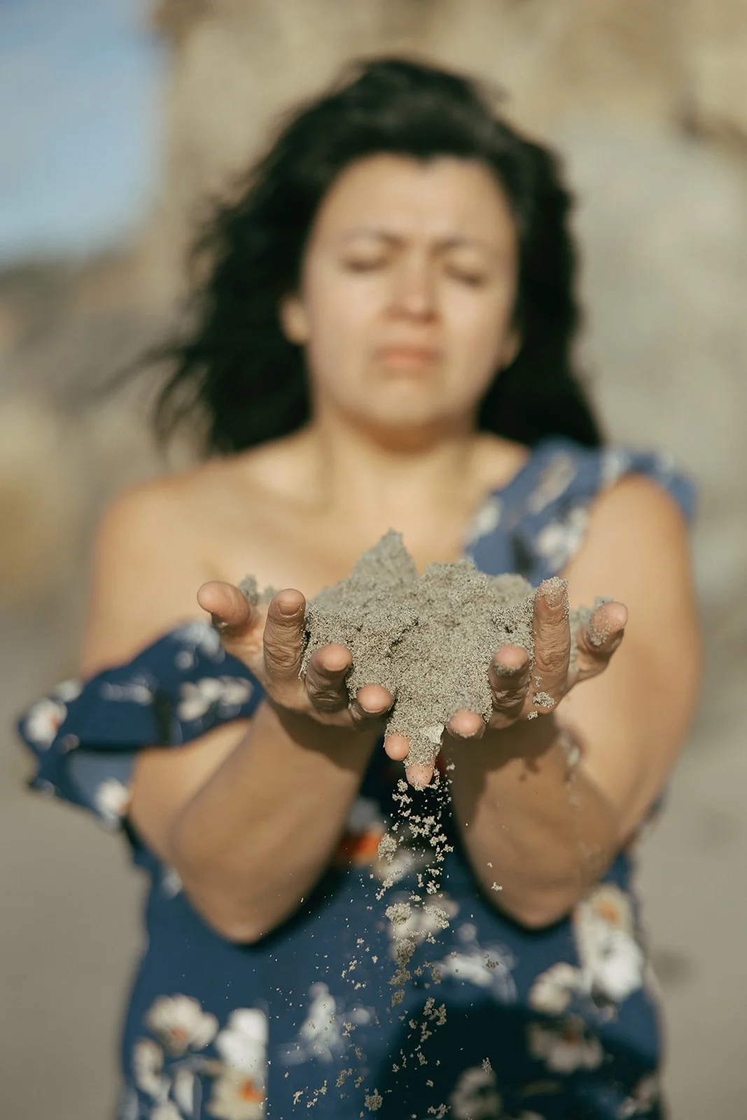 A woman with dark hair and a blue floral dress holds sand in her hands, with some falling from her hands. Brand and lifestyle photography for Connie Neyra and yoga practitioner by Rhianon Brown Photography.