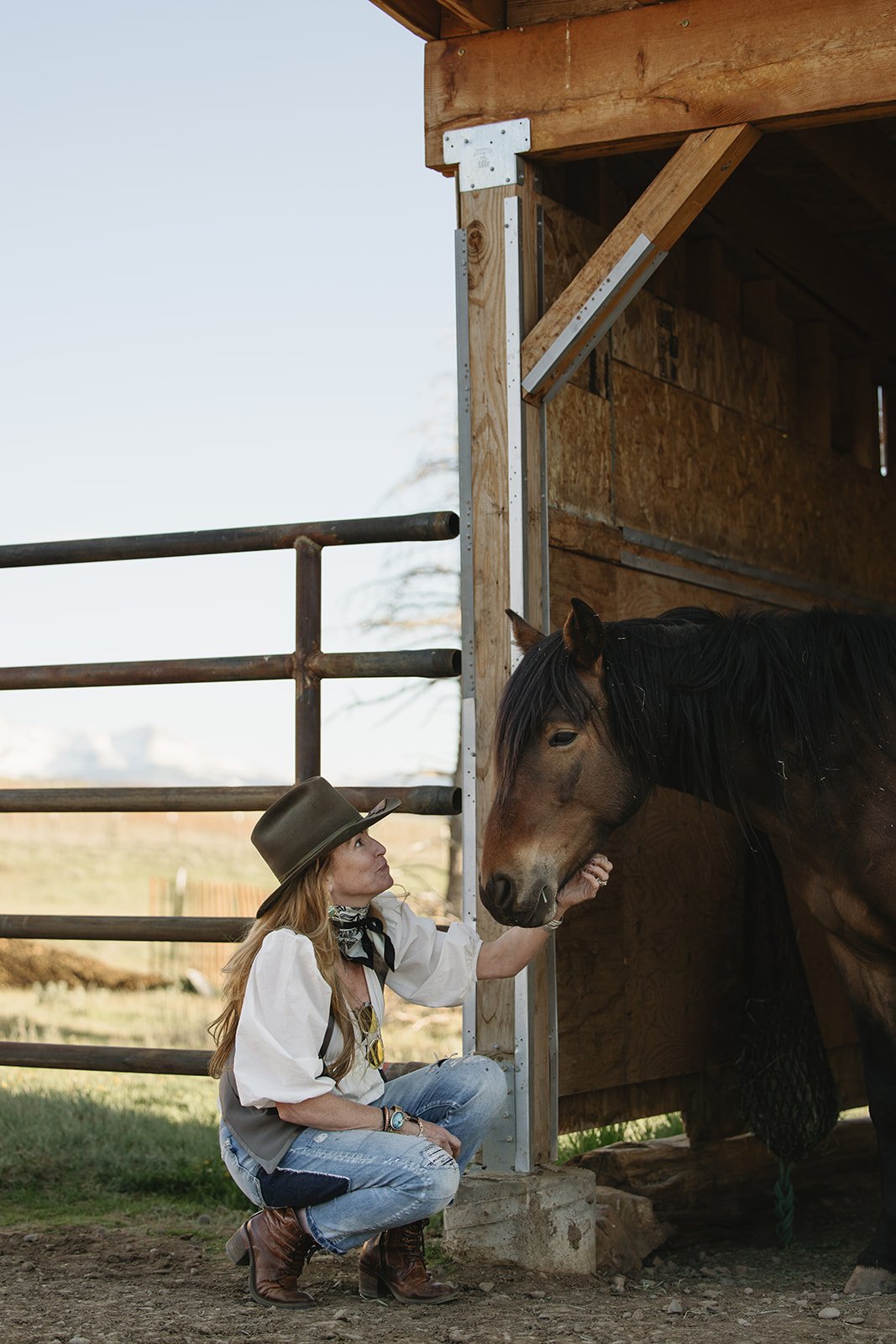 A woman with long red hair, wearing a cowboy hat, a white blouse, and jeans, crouches down on one knee and gently touches the head of a brown horse inside a wooden stable. Rhianon Brown Photography's brand photoshoot for Grace Reins.