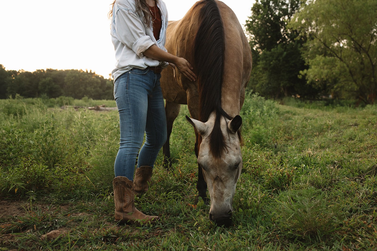 A woman in jeans and cowboy boots feeding a light brown horse in a green field with trees in the background.  Brand and lifestyle photography for Sacred Align Equine by Rhianon Brown Photography.