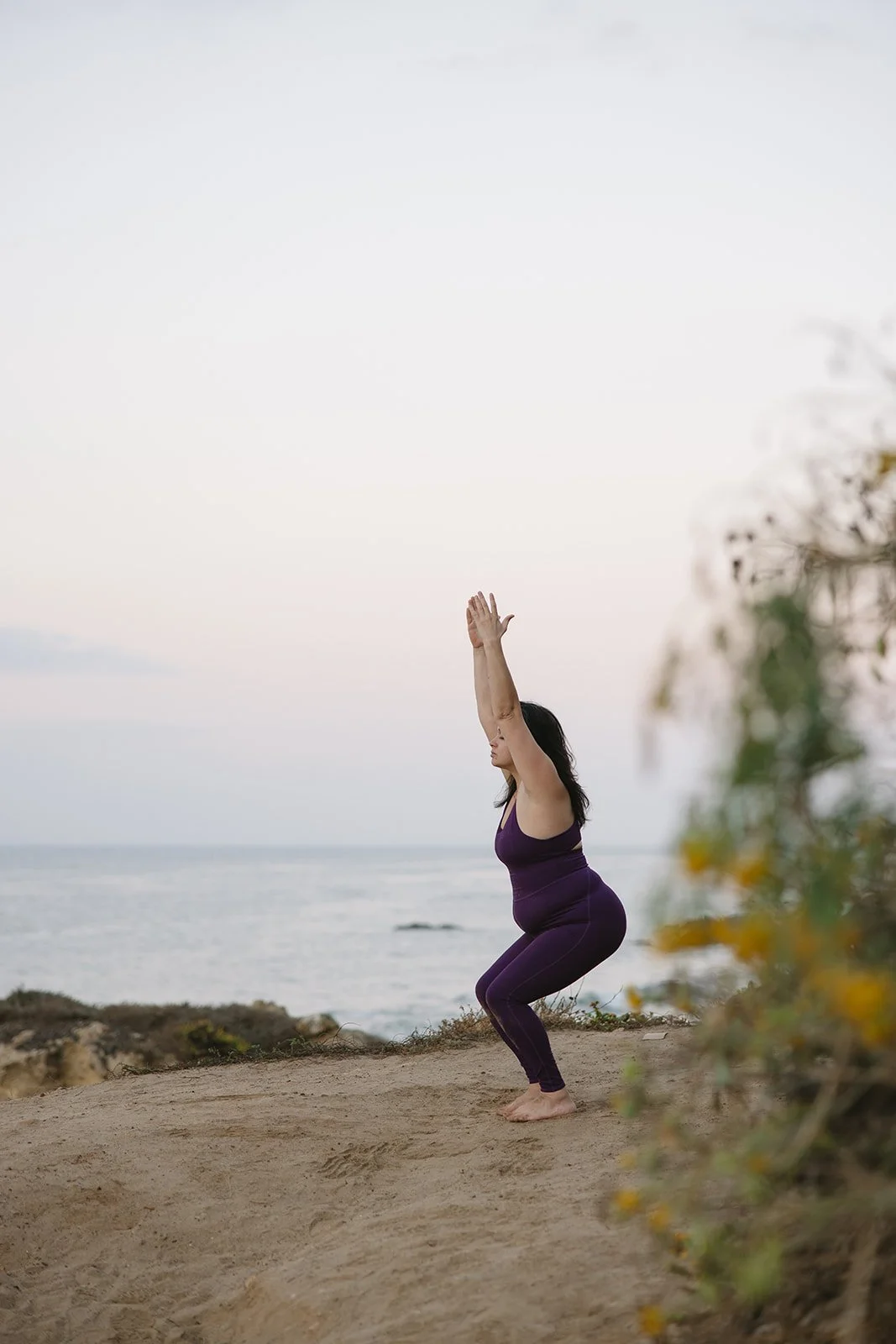Woman practicing yoga outdoors on a dirt path near the beach, raising her arms overhead in a squat position during sunset. Brand and lifestyle photography for Connie Neyra and yoga practitioner by Rhianon Brown Photography.