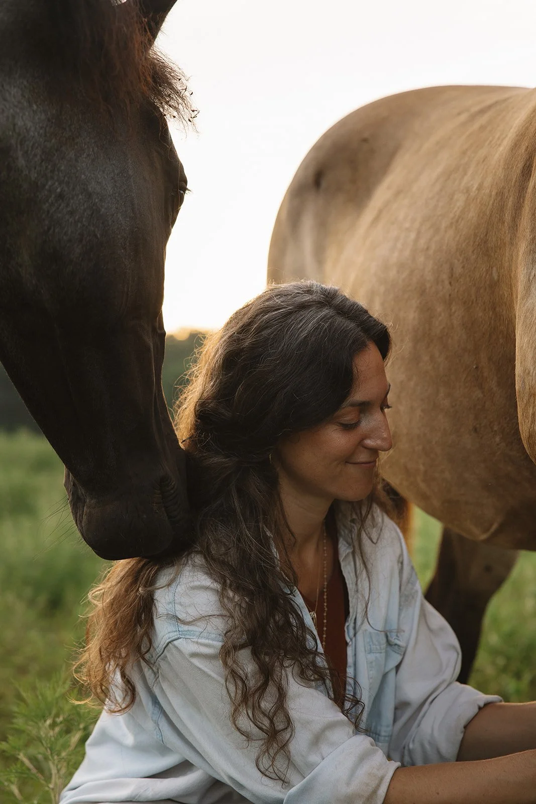 A woman with long, curly hair is sitting outdoors in a grassy field, smiling with her eyes closed, surrounded by three large horses during sunset. Brand and lifestyle photography for Sacred Align Equine by Rhianon Brown Photography.