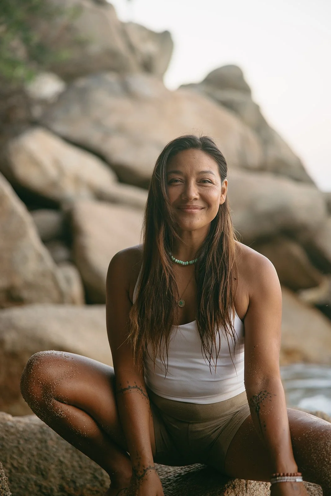 A woman with long brown hair sitting on rocks at the beach, smiling, wearing a white tank top, shorts, and jewelry, with sand on her knees and arms. Brand and retreat photography for Nikki Nie by Rhianon Brown Photography.