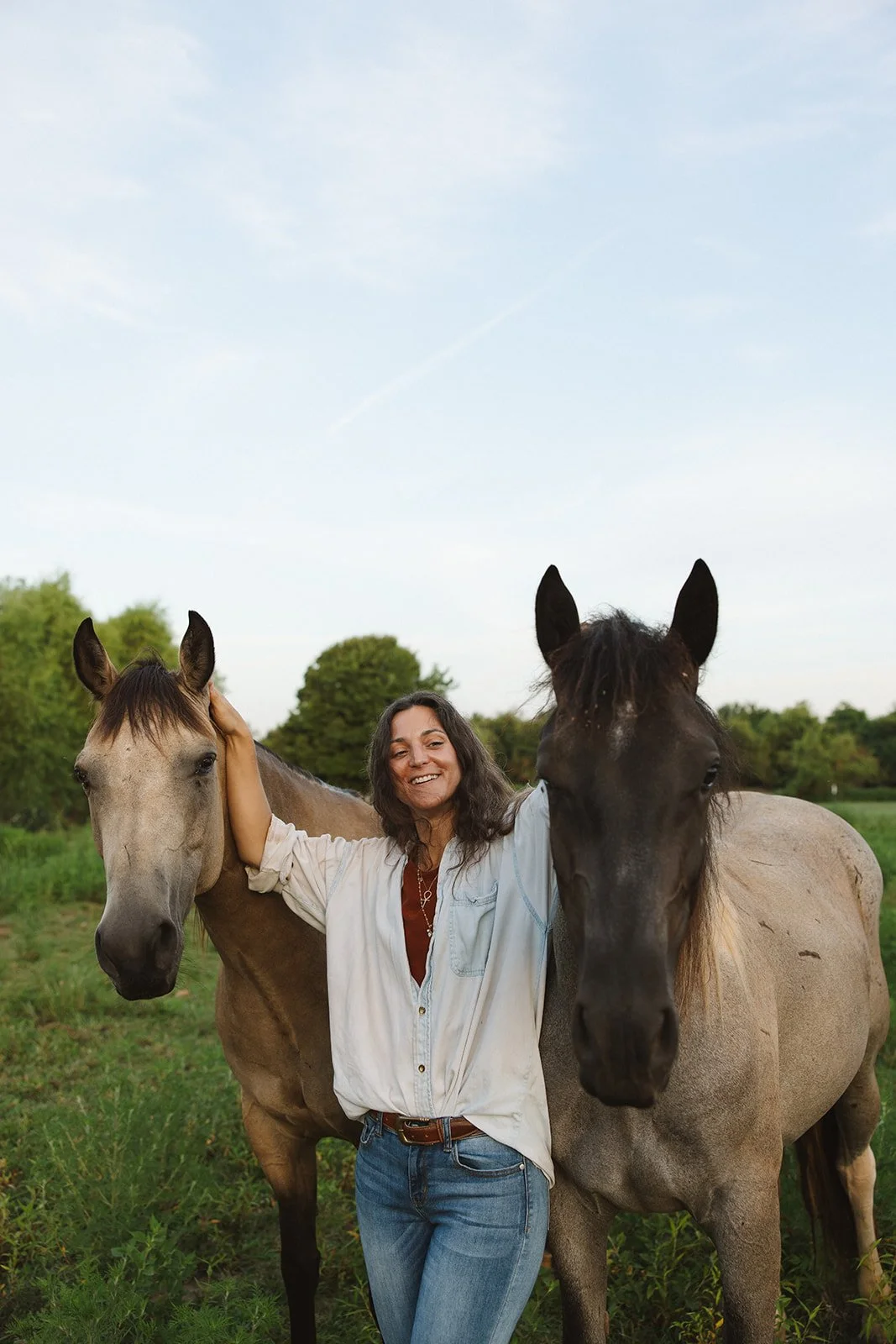 A woman standing between two horses, smiling and touching their heads, in a green outdoor setting with trees and a clear sky.  Brand and lifestyle photography for Sacred Align Equine by Rhianon Brown Photography.