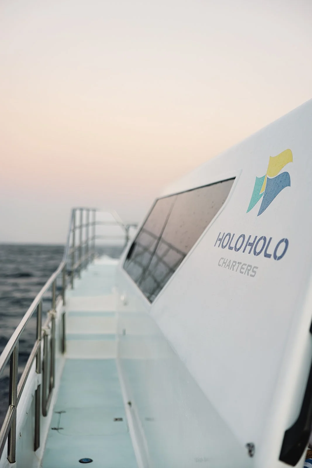 Close-up photo of a boat with the logo and text 'HOLOHOLO CHARTERS' on its side, on the ocean during sunset, with the horizon and sky in the background. Brand and travel photography session for Holo Holo Charters in Kauai, Hawaii by Rhianon Brown Pho