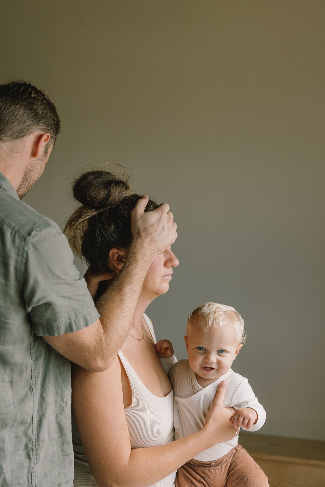 A man is fixing a woman's hair while she holds a young child with blonde hair and blue eyes, who is smiling and looks playful.