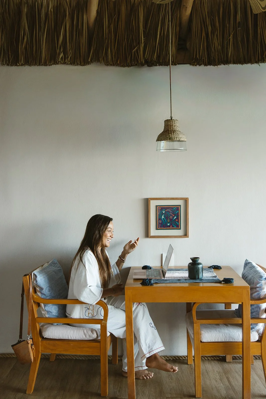 A woman sitting at a wooden dining table, smiling and talking on a cell phone. She is wearing a white robe and sitting on a cushioned chair with pillows. Brand and retreat photography for Nikki Nie by Rhianon Brown Photography.