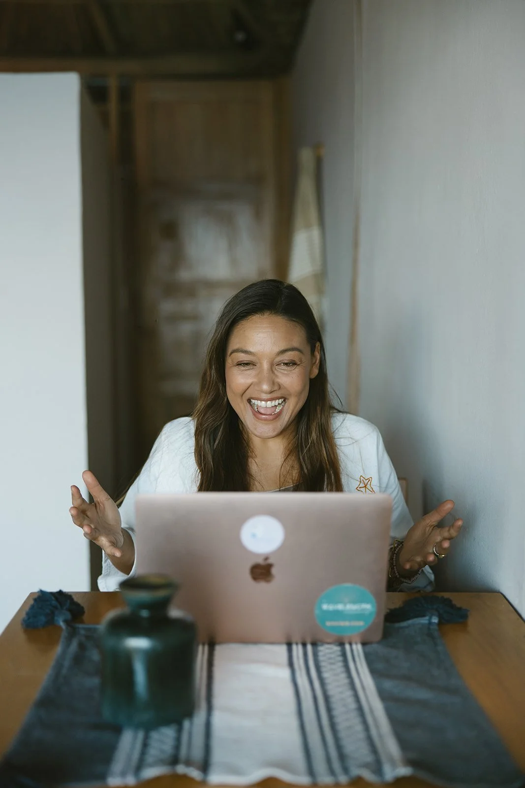 A woman sitting at a dining table using a laptop, smiling and gesturing with her hands. Brand and retreat photography for Nikki Nie by Rhianon Brown Photography.