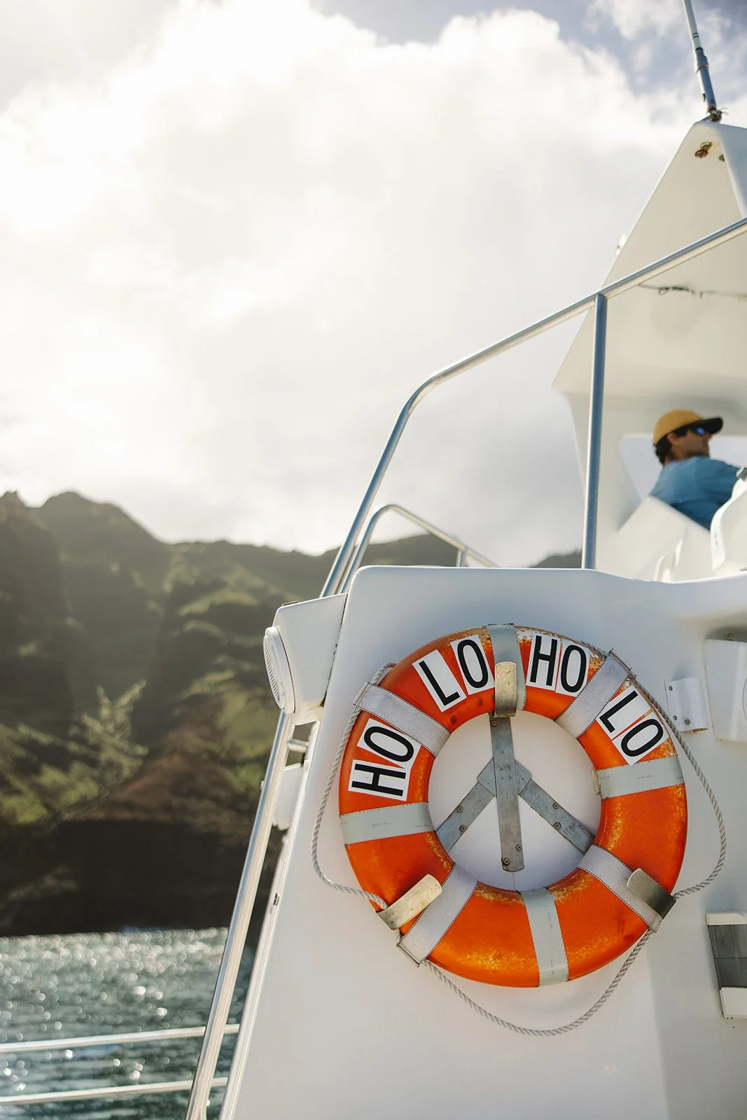Close-up of a life preserver with the word "ALOHA" repeated around it mounted on the side of a boat, with a scenic coastline and sky in the background.