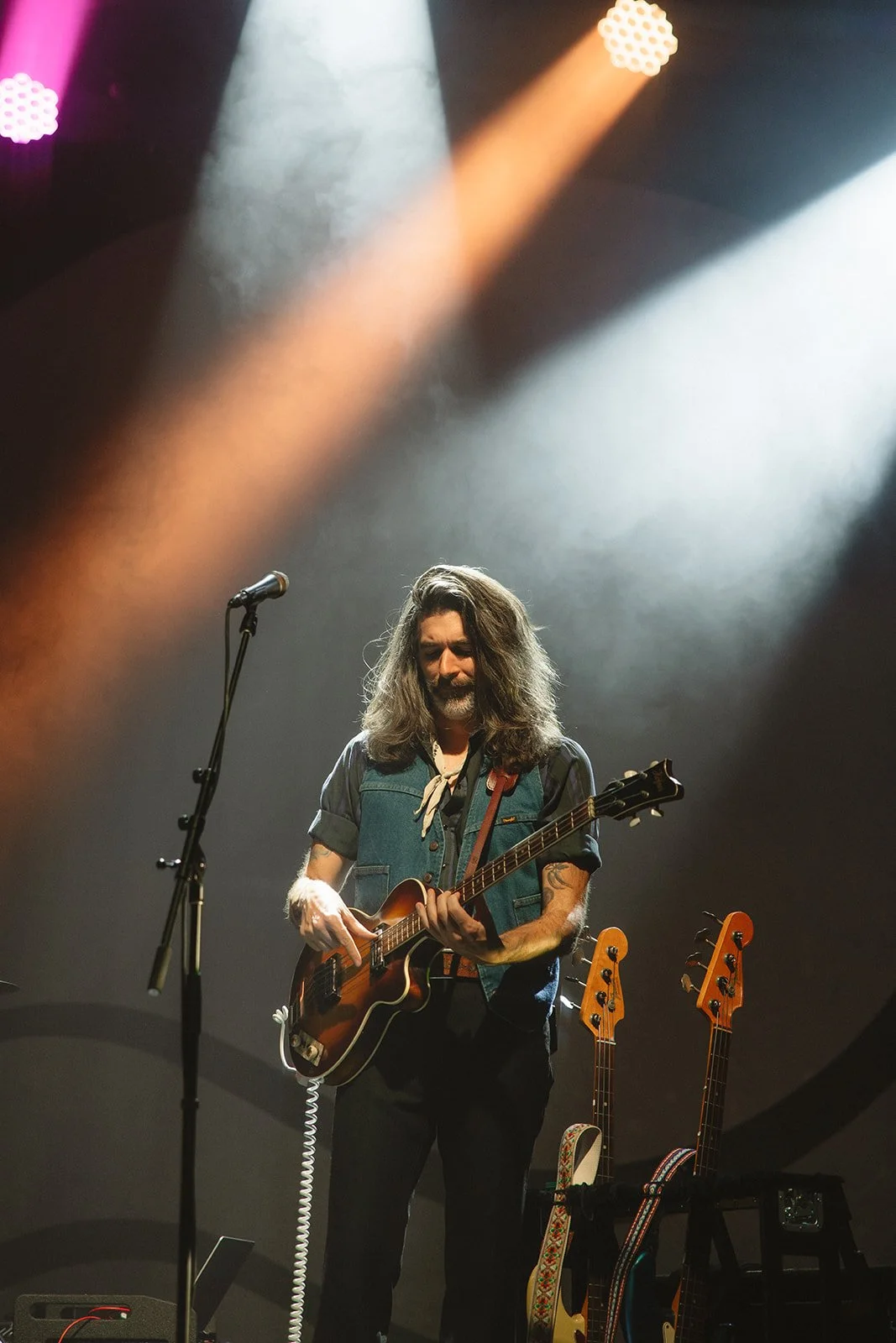 A man with long hair and a beard playing an electric guitar on stage, illuminated by stage lights, with two additional guitars in the background. Aly and Aj concert event photographed by Rhianon Brown Photography.