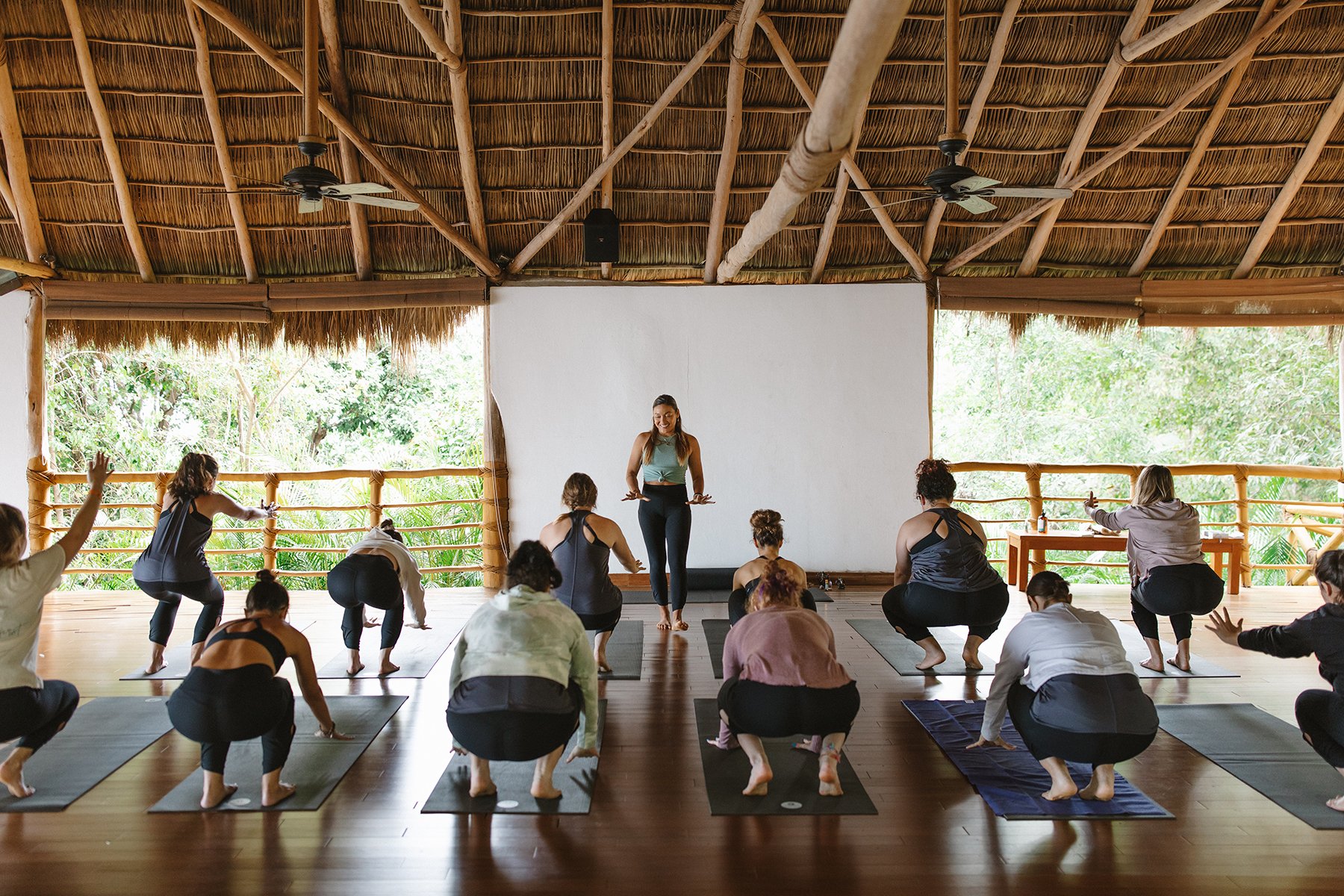 A woman leading a yoga class in a bamboo and thatch open-air studio surrounded by greenery, with students practicing yoga poses on mats. Brand and retreat photography for Nikki Nie by Rhianon Brown Photography.