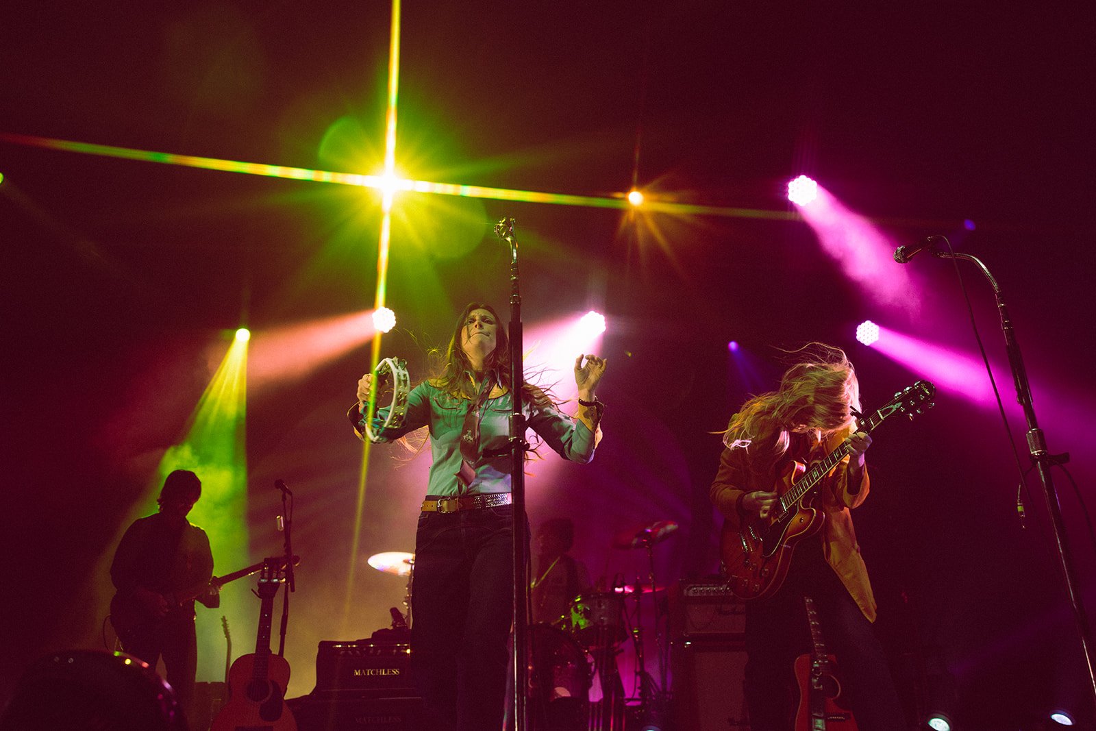 Musicians performing on stage with colorful lights, including a woman singing with a tambourine, a guitarist, and a bass player, during a concert. Aly and Aj concert event photographed by Rhianon Brown Photography.
