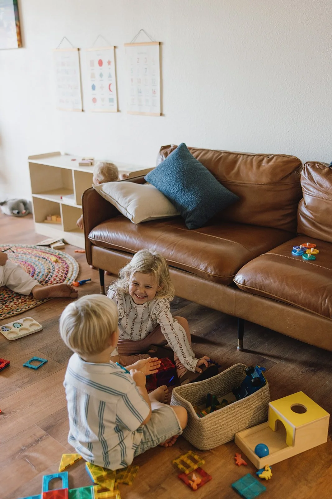 Children playing with toys on the floor of a living room, with a brown leather couch, a white bookshelf, and children's artwork on the wall in the background. Brand and lifestyle photography session for Abundant Life Chiropractics in Maui, Hawaii by
