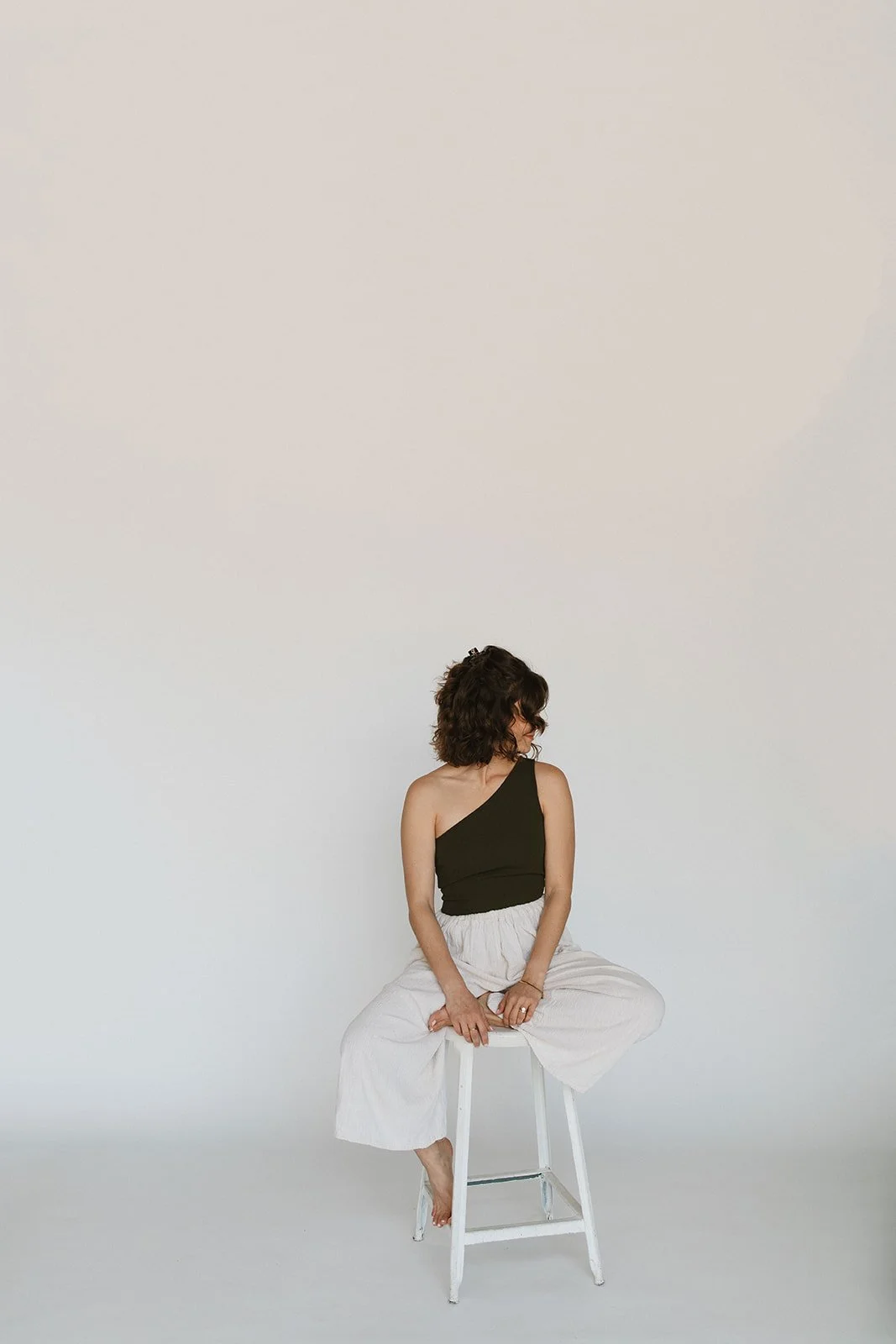 Woman sitting on a white stool against a plain white wall, wearing a black one-shoulder top and white wide-leg pants, looking to her left. Branding photoshoot for Motherland Wellness by Rhianon Brown Photography in Springfield, Missouri.