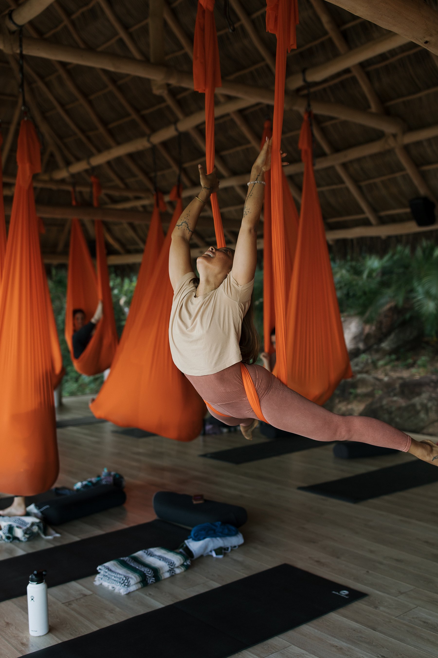 A woman practicing aerial yoga on orange fabric swings in a wooden, open-air yoga studio with a thatched roof. Brand and retreat photography for Nikki Nie by Rhianon Brown Photography.