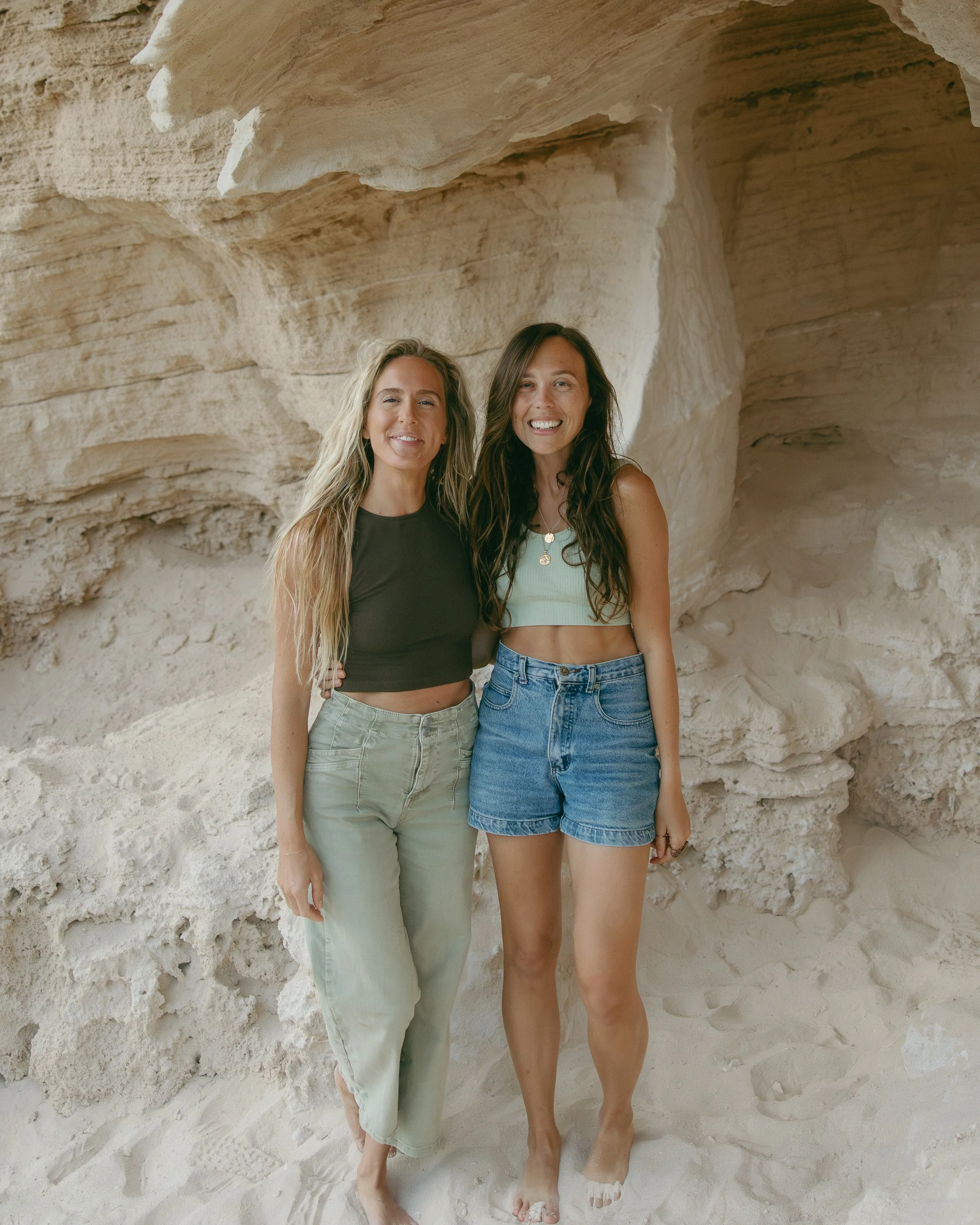 Two women with long hair standing side by side on a sandy surface with a rocky formation behind them. Bekah Cooper of Forma Made and Rhianon Brown of Rhianon Brown Photography