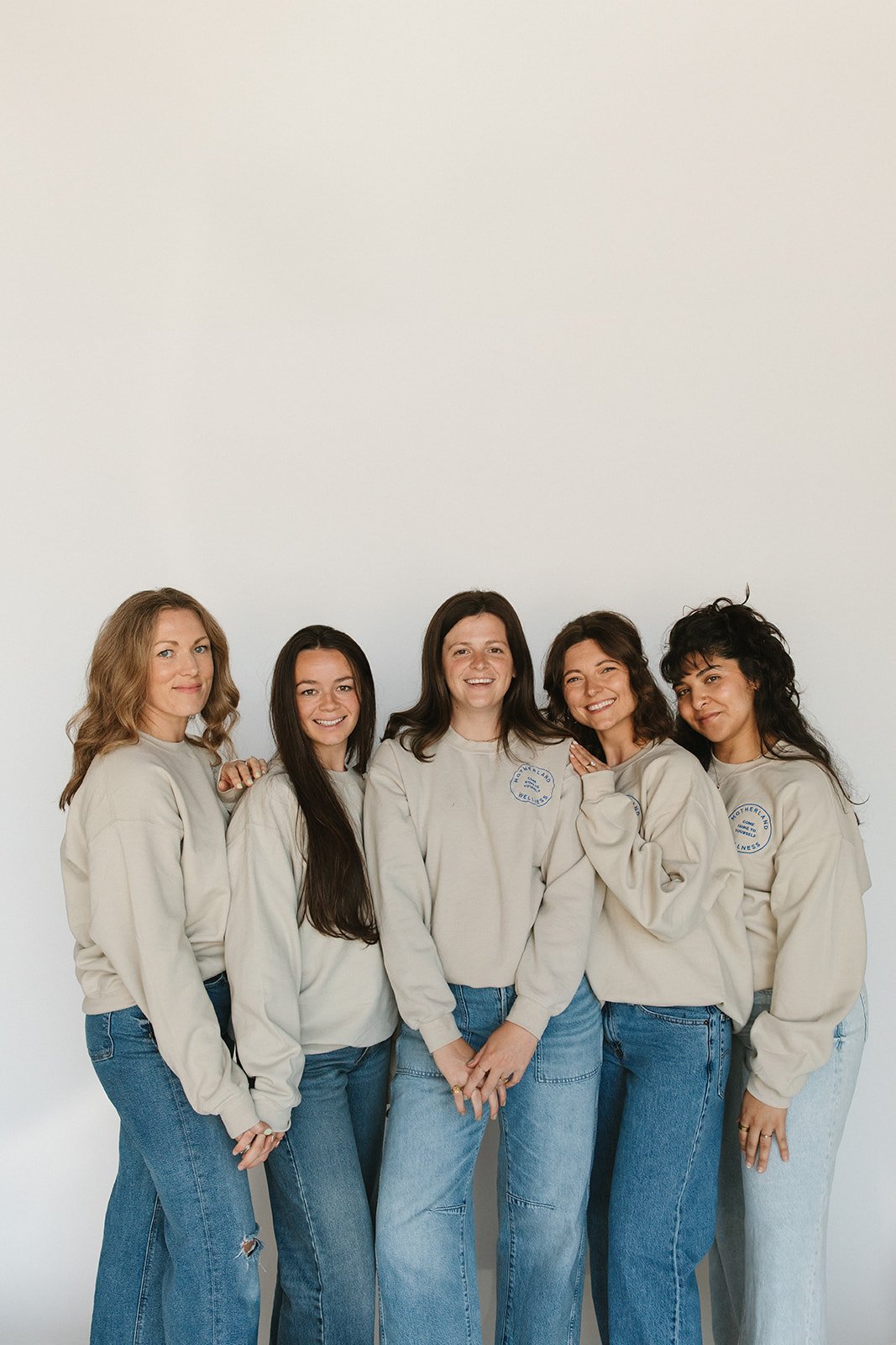 Group of five women standing together, smiling, wearing matching beige sweatshirts and jeans against a plain white background. Branding photoshoot for Motherland Wellness by Rhianon Brown Photography in Springfield, Missouri.