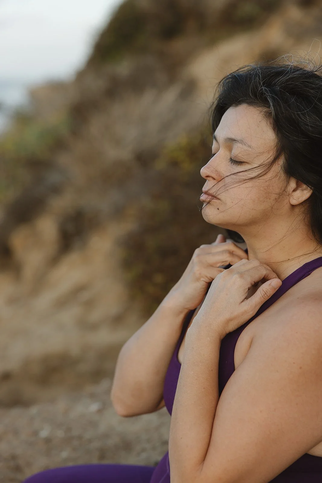 A woman sitting outdoors on a rocky, earthy terrain with her eyes closed and hands resting on her chest and shoulder, with wind gently blowing her hair. Brand and lifestyle photography for Connie Neyra and yoga practitioner by Rhianon Brown