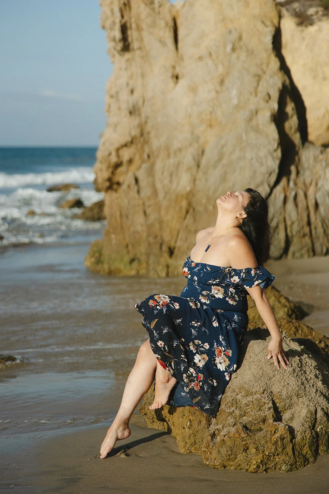 A woman sitting on a rock near the beach with cliffs in the background, wearing a floral off-shoulder dress and enjoying the sunlight. Brand and lifestyle photography for Connie Neyra and yoga practitioner by Rhianon Brown Photography.