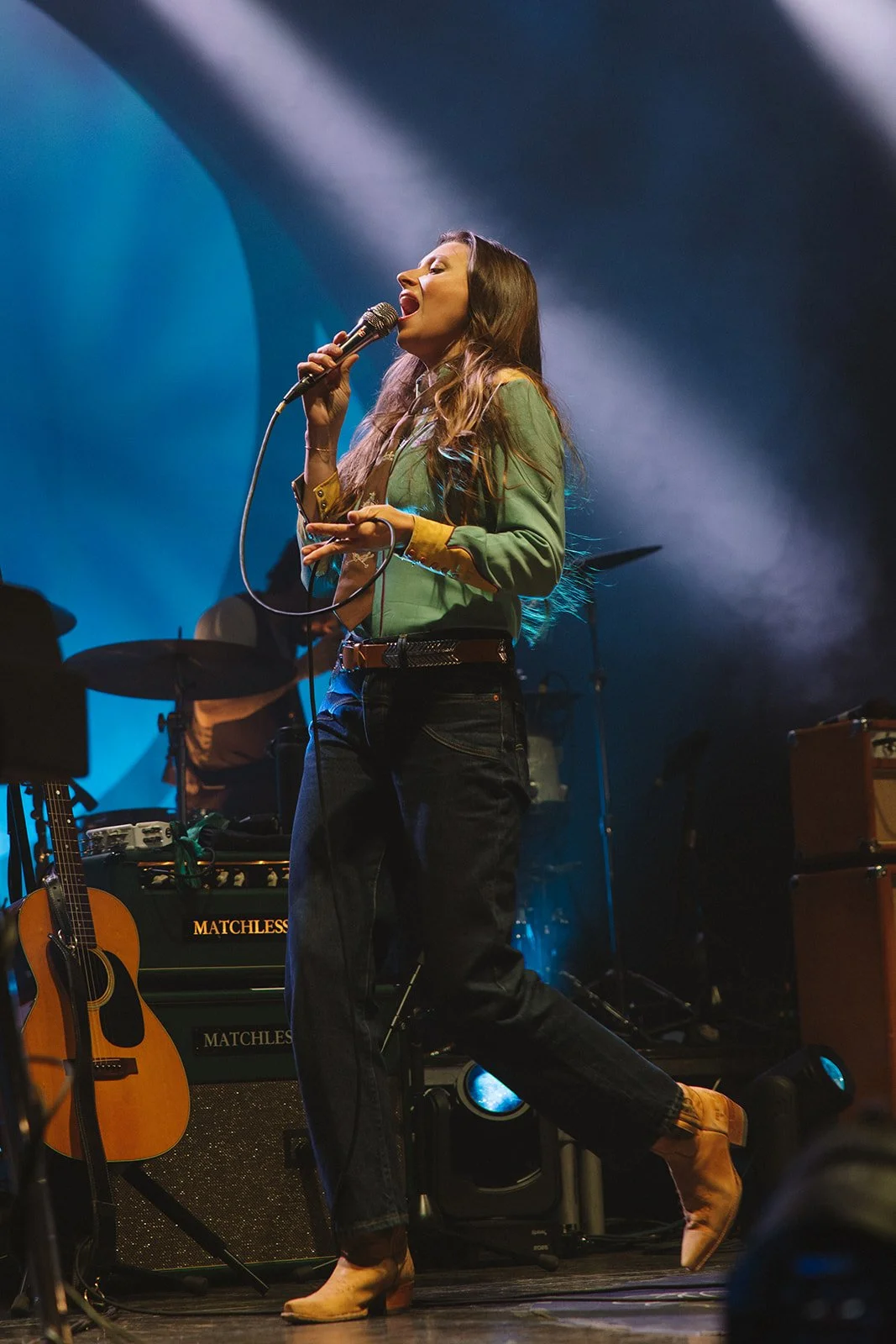 A woman singing into a microphone on stage with musical equipment, including a guitar and amplifiers, behind her. Aly and Aj concert event photographed by Rhianon Brown Photography.