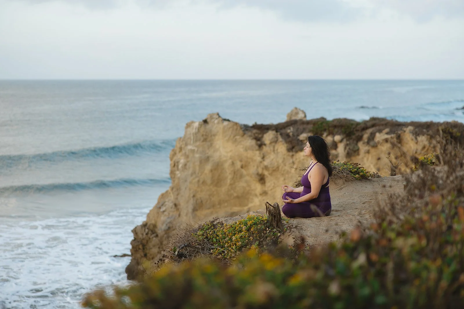 Woman practicing yoga on a rocky cliff overlooking the ocean, wearing a purple sports top and leggings, with waves crashing below. Brand and lifestyle photography for Connie Neyra and yoga practitioner by Rhianon Brown Photography.