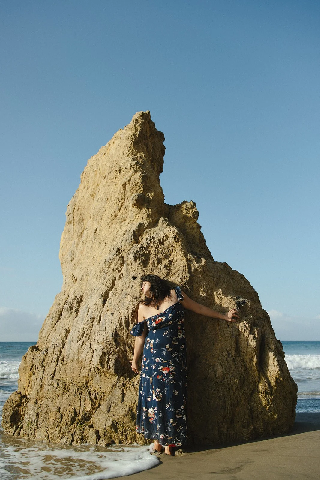 A woman in a floral dress standing on a sandy beach, touching a large rock formation with the ocean and blue sky in the background. Brand and lifestyle photography for Connie Neyra and yoga practitioner by Rhianon Brown Photography.