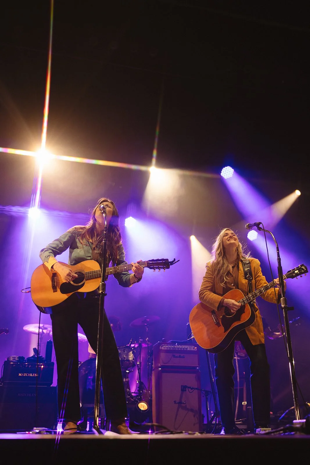 Two women playing acoustic guitars and singing on stage during a live music performance, with colorful stage lights shining overhead. Aly and Aj concert event photographed by Rhianon Brown Photography.
