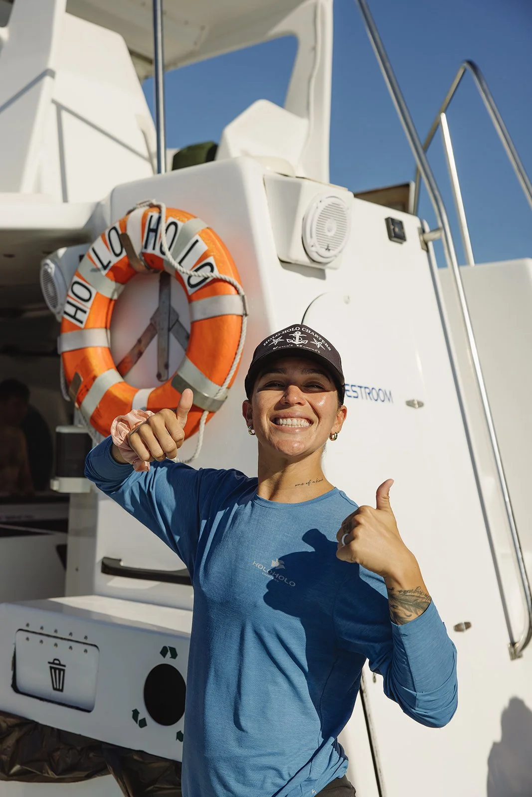 A woman smiling and giving a thumbs up in front of a boat with a life ring and a clock on it, wearing a black cap and a blue long-sleeve shirt. Brand and travel photography session for Holo Holo Charters in Kauai, Hawaii by Rhianon Brown Photography.