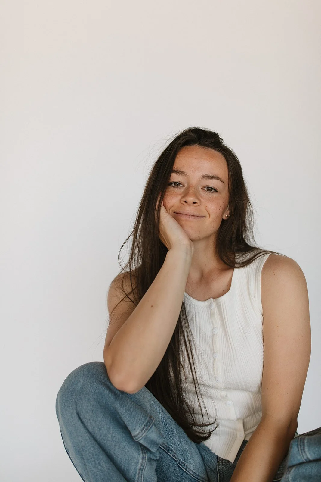 Portrait of a young woman with long dark hair, sitting against a plain white wall, smiling softly, wearing a sleeveless white top and blue jeans. Branding photoshoot for Motherland Wellness by Rhianon Brown Photography in Springfield, Missouri.