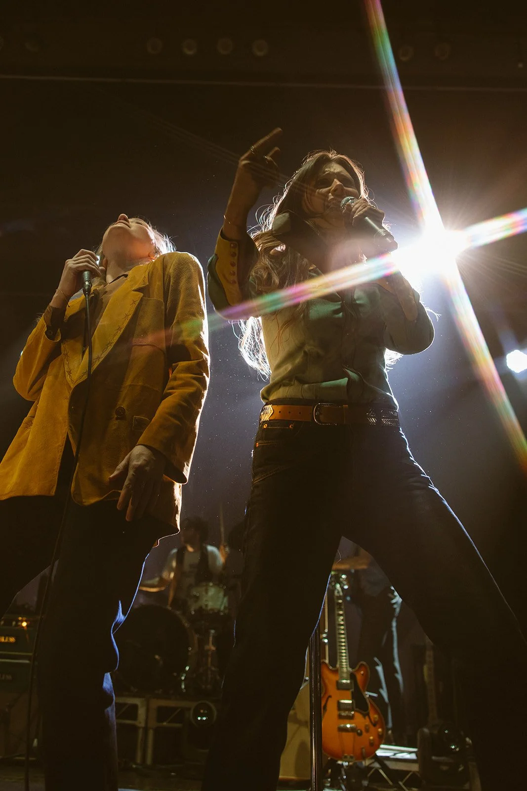 Two female singers performing on stage under bright lights, with one wearing a yellow jacket and the other wearing a green shirt, surrounded by musical instruments and band equipment. Aly and Aj concert event photographed by Rhianon Brown Photography