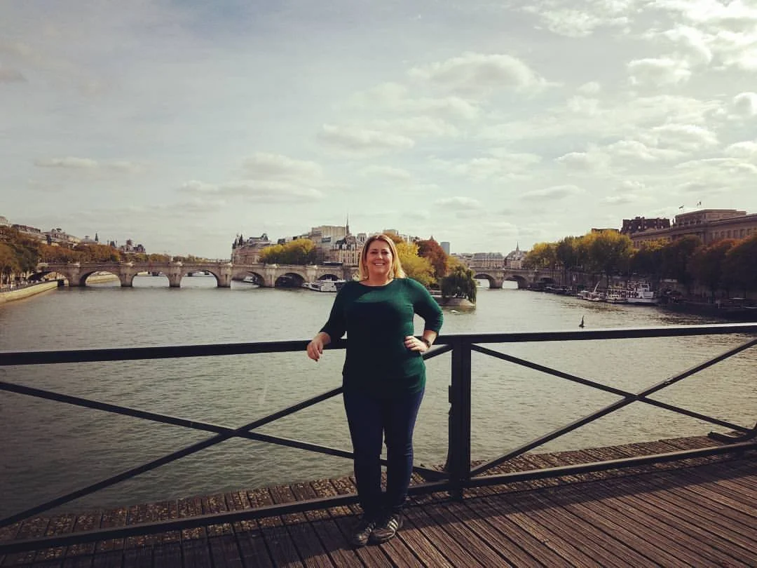 Jen Cease, CMP standing on a bridge in Paris on the Seine with a cityscapein the background, under a partly cloudy sky.