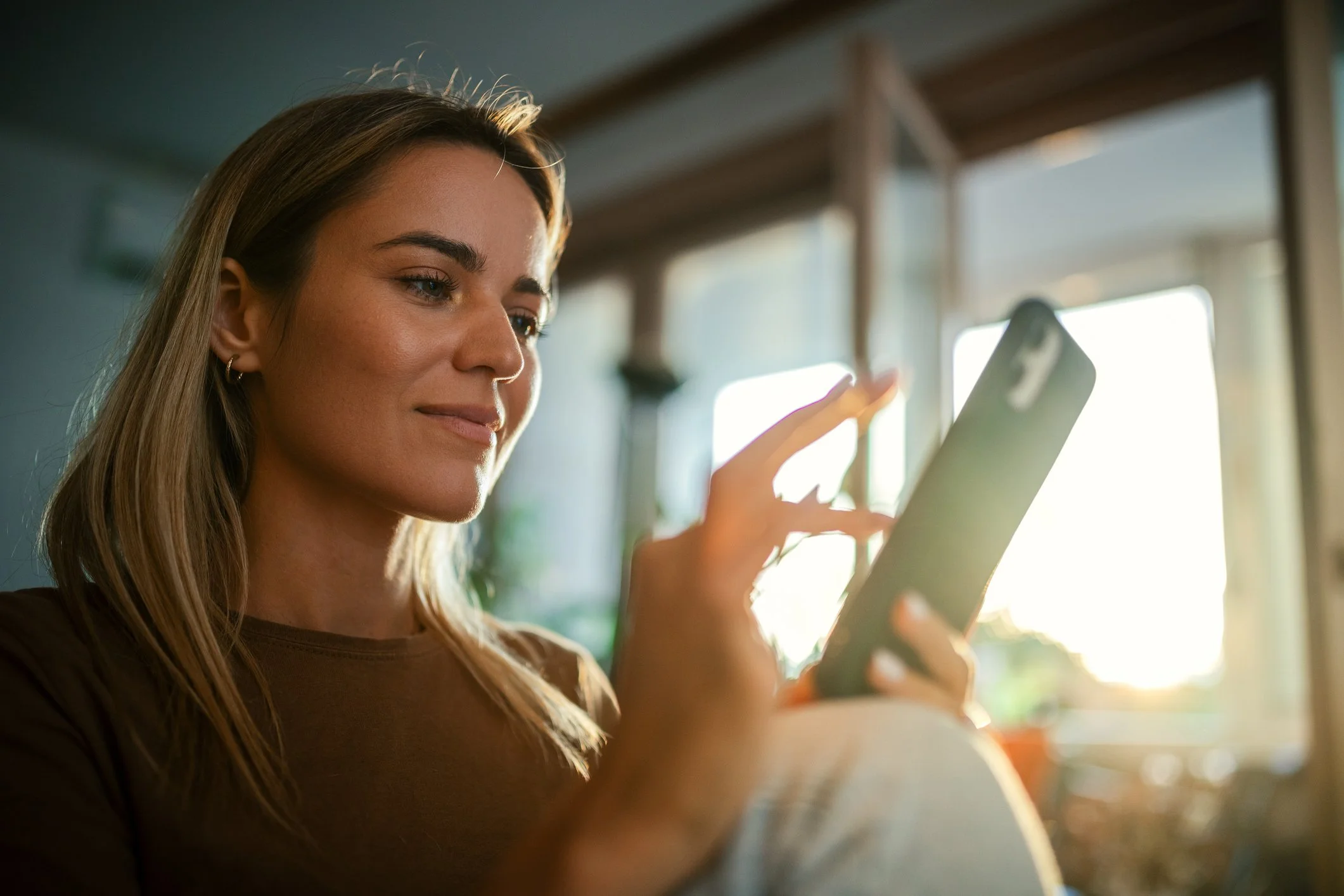 A young woman with long brown hair and hoop earrings looking at her phone in a cozy, sunlit room.